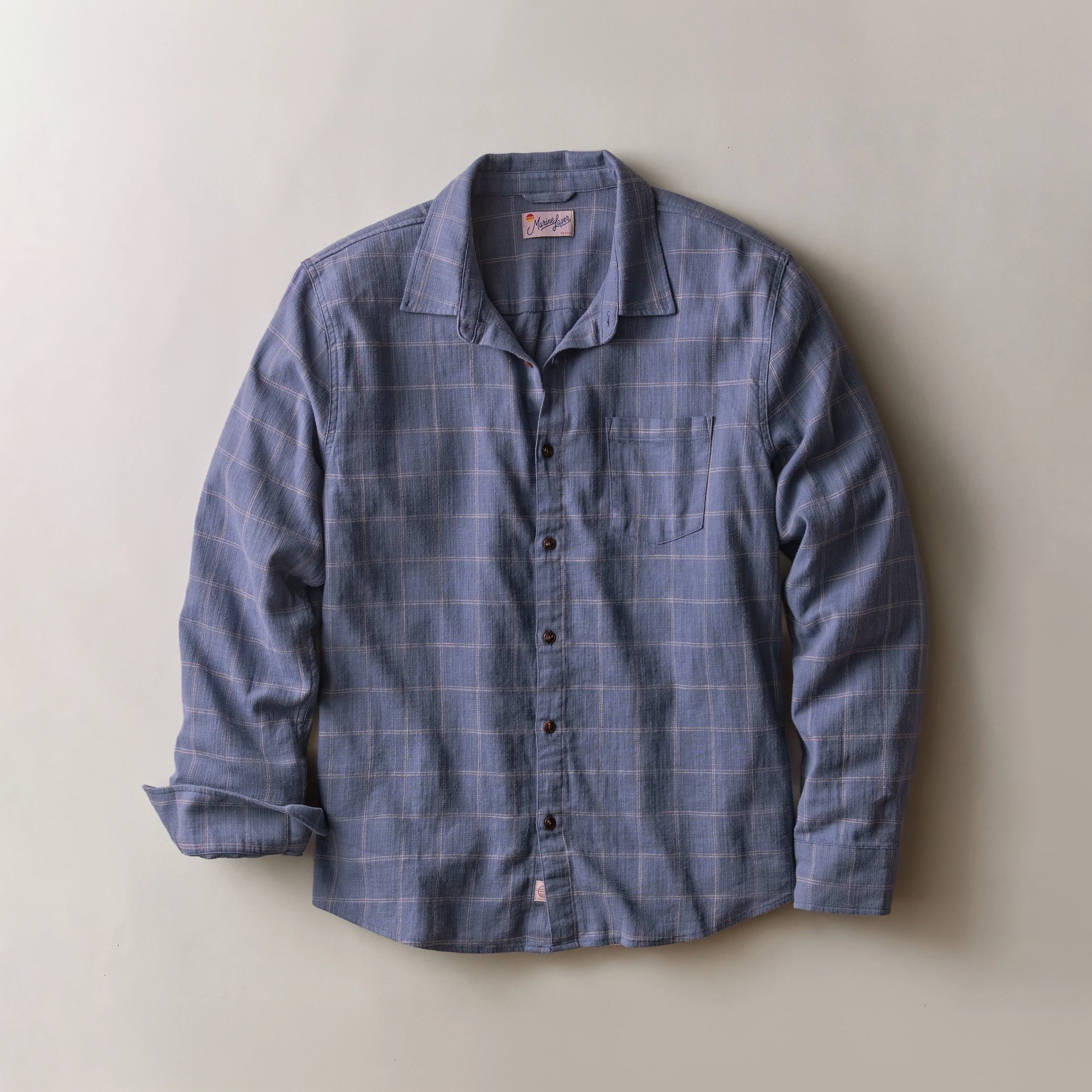 A long-sleeve button-up shirt in a dusty blue plaid pattern, laid flat against a neutral beige background.