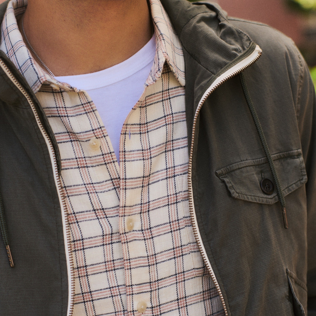 Close-up of a person wearing an olive green zip-up jacket over a cream plaid button-up shirt and a white t-shirt.