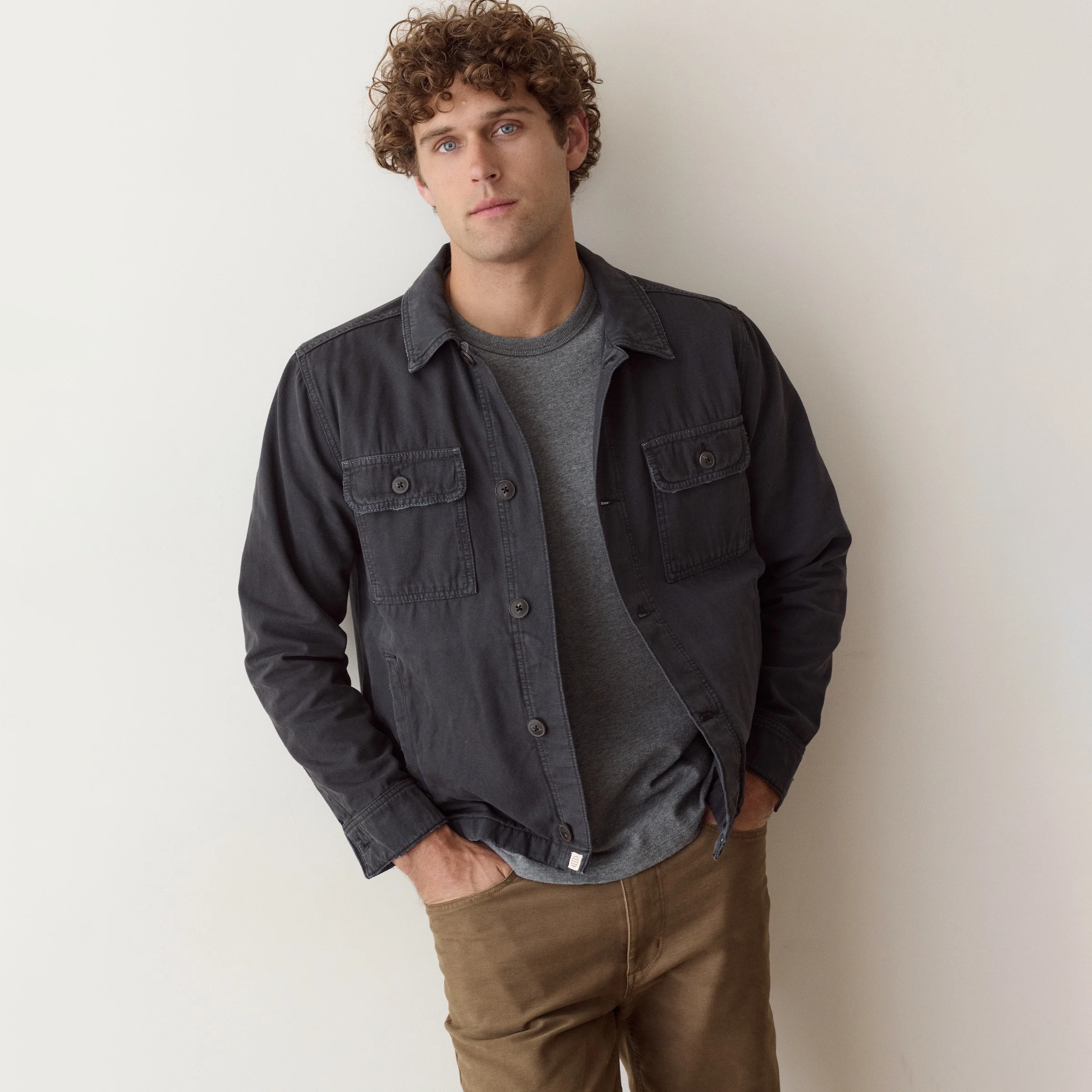 A man wearing a dark charcoal gray button-up shirt jacket over a gray t-shirt and brown pants, standing against a plain off-white wall.