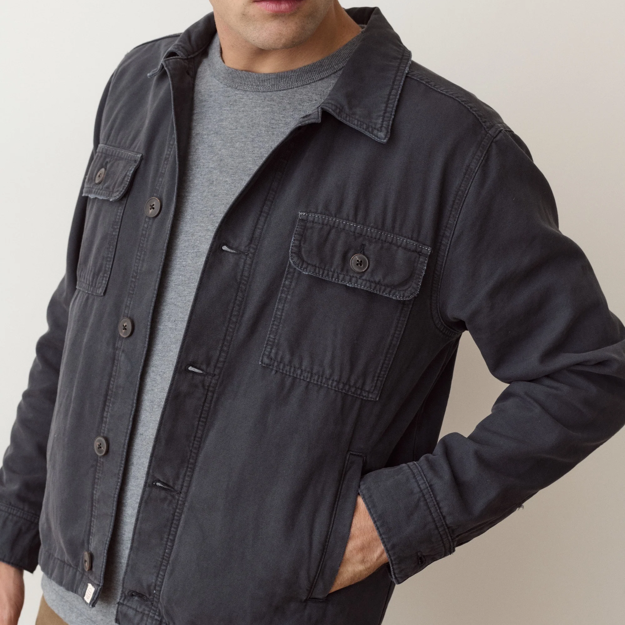 A man wearing a dark charcoal gray utility jacket over a gray crewneck t-shirt, shown from the chest down against a light beige background.