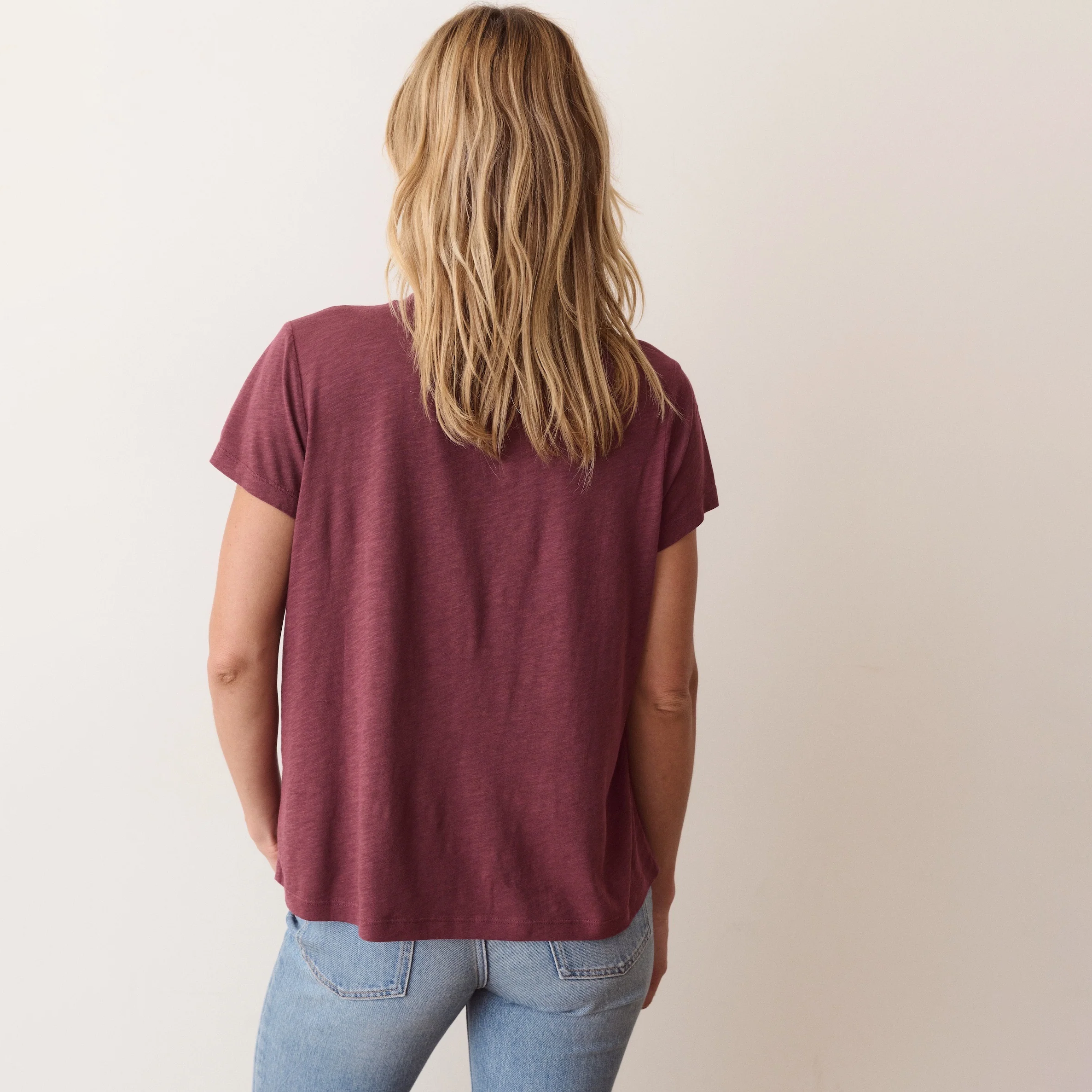 A woman with blonde hair is shown from behind wearing a heathered maroon short-sleeve t-shirt and light blue jeans against a plain off-white background.