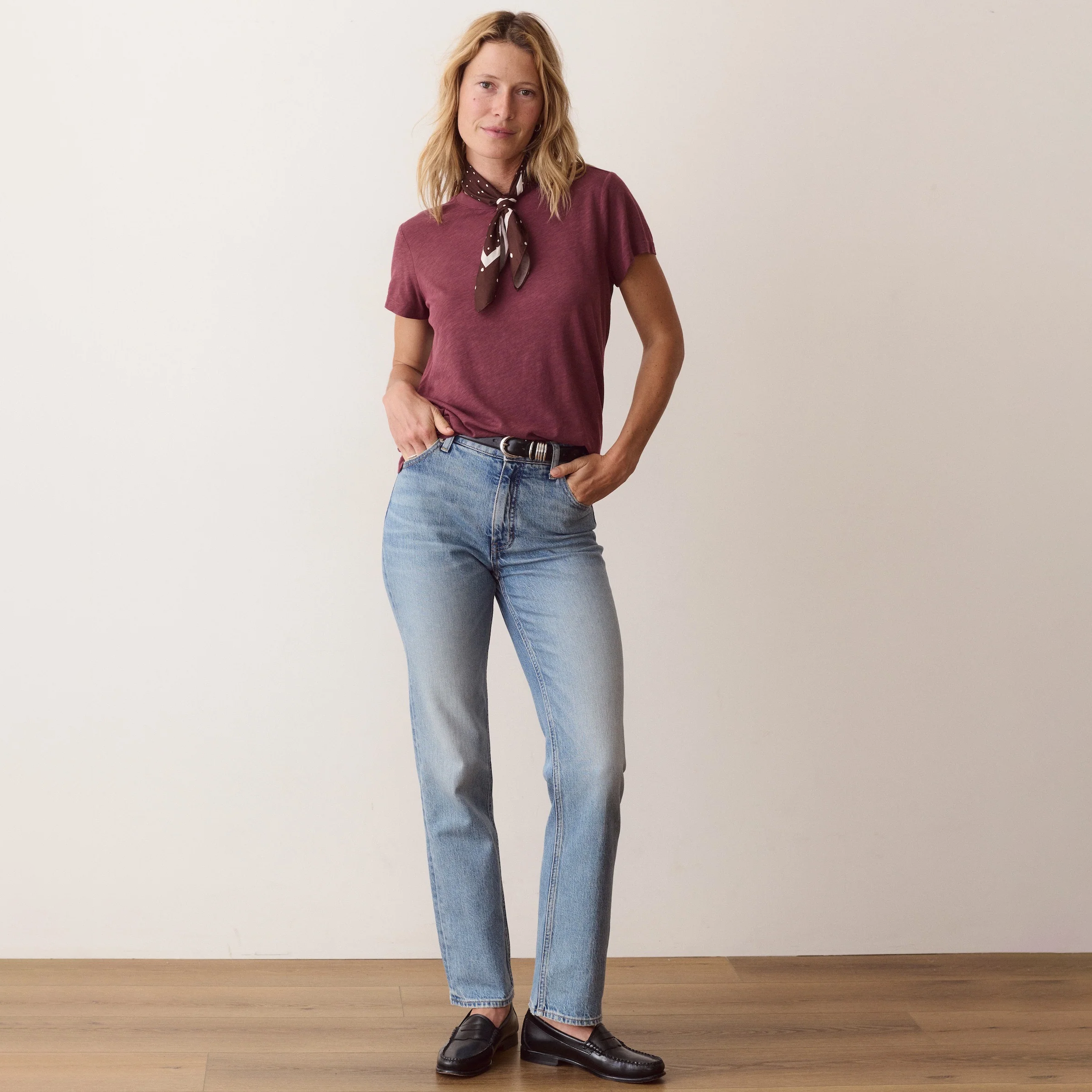 A woman stands in a studio wearing a short-sleeve burgundy shirt with a dark patterned neck scarf, light-wash blue jeans, a black belt, and dark brown leather loafers, against a plain off-white wall and light wood floor.
