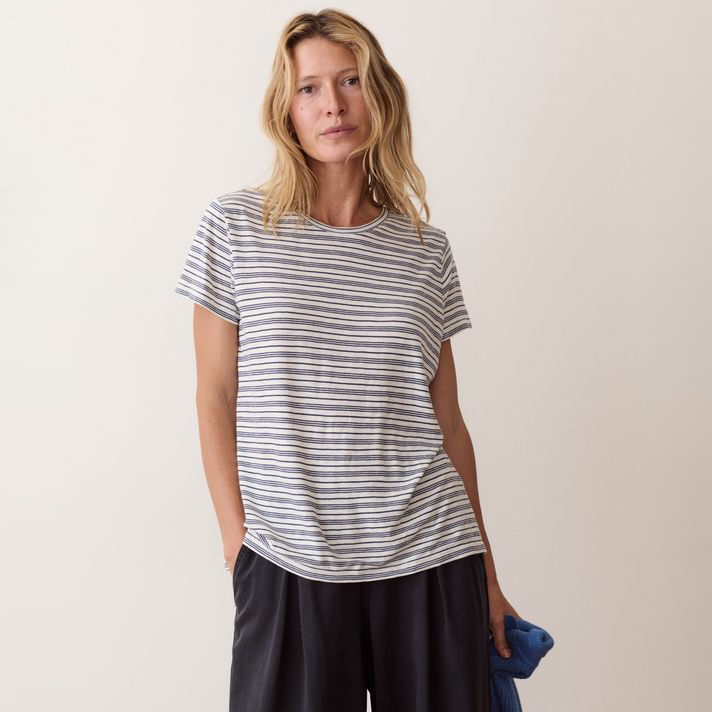 A woman modeling a short-sleeve crewneck t-shirt with thin navy and white horizontal stripes, paired with dark pants, against a plain light beige studio background.