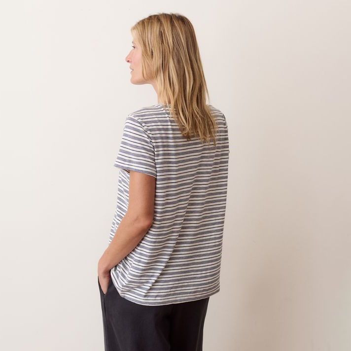 A woman with blonde hair is shown from the back wearing a short-sleeve t-shirt with thin navy and white horizontal stripes, paired with dark pants, against a plain beige background.