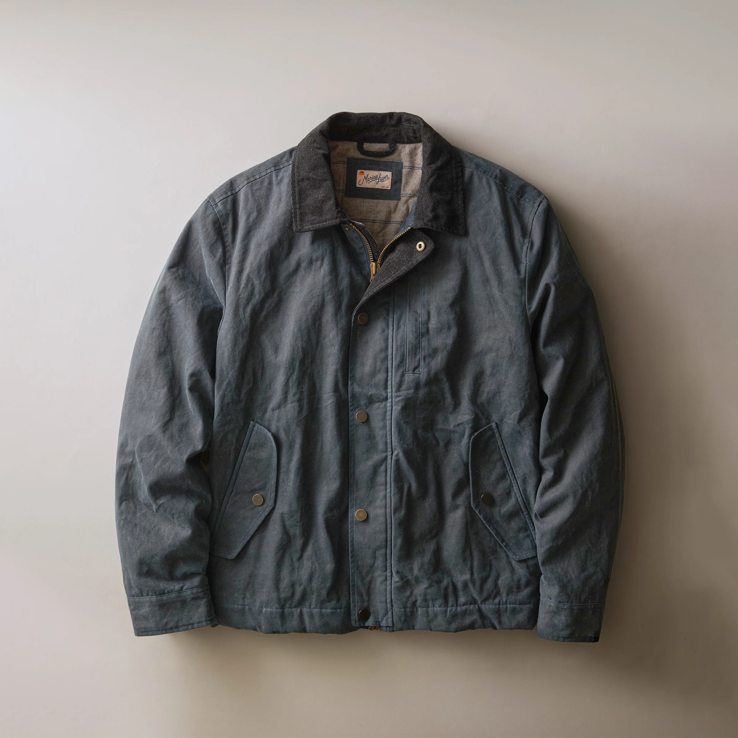 A dark slate blue waxed canvas utility jacket with a black corduroy collar, brown buttons, and a zippered front, displayed flat against a neutral beige background.