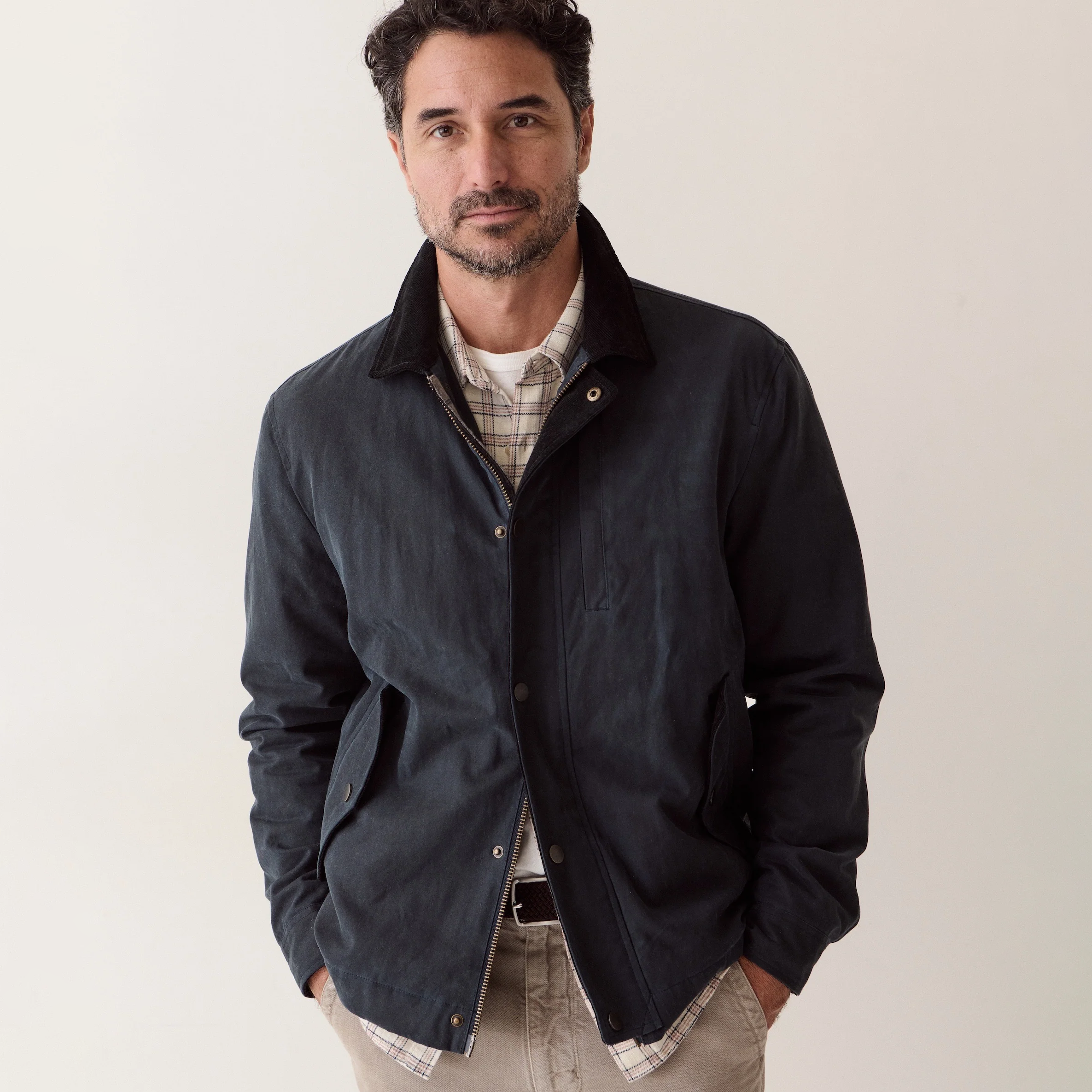 A man wearing a dark navy waxed cotton jacket over a beige plaid shirt and khaki pants, standing against a plain off-white background.