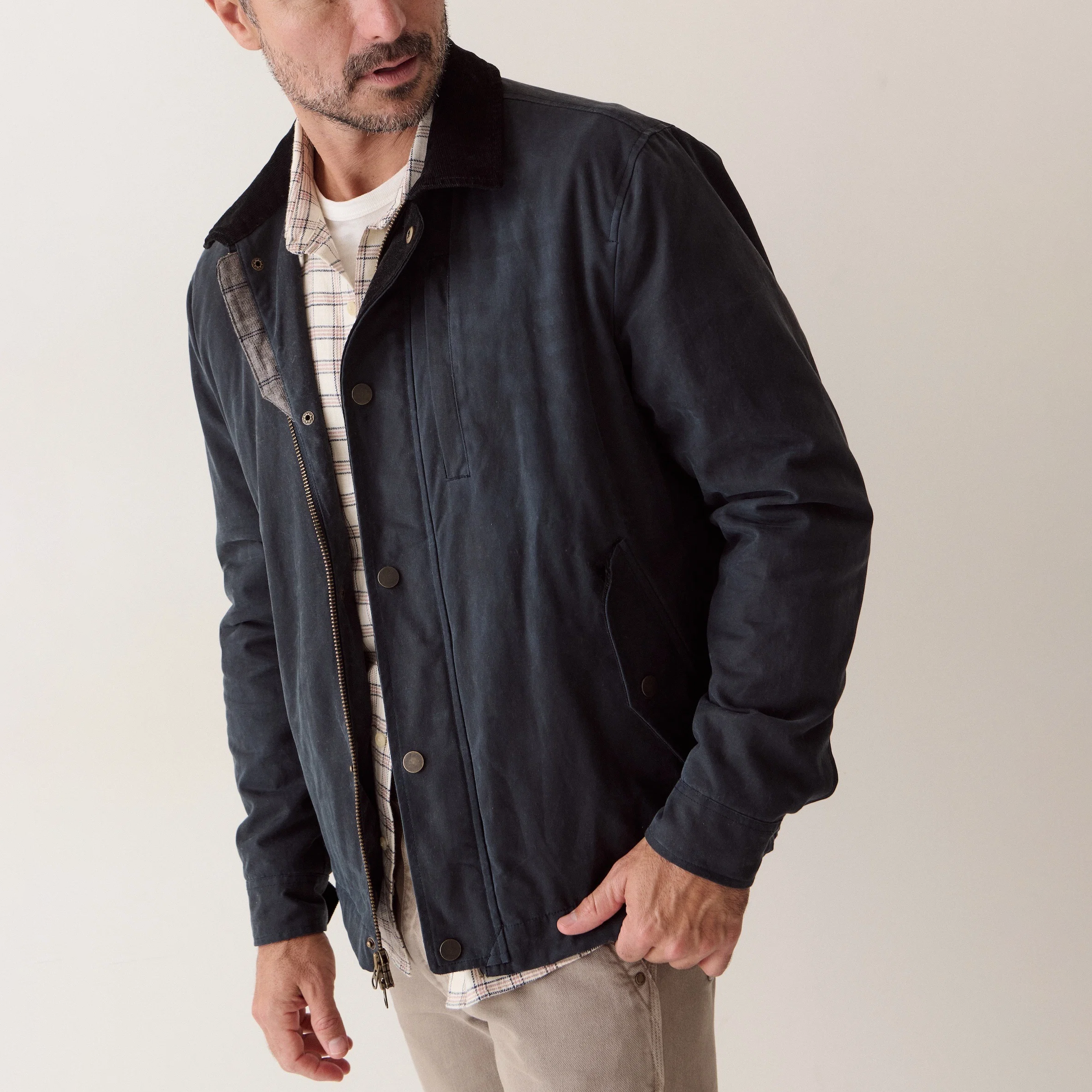 A man wearing a dark navy waxed cotton jacket over a beige and white plaid shirt, paired with light beige trousers, photographed against a neutral off-white background.