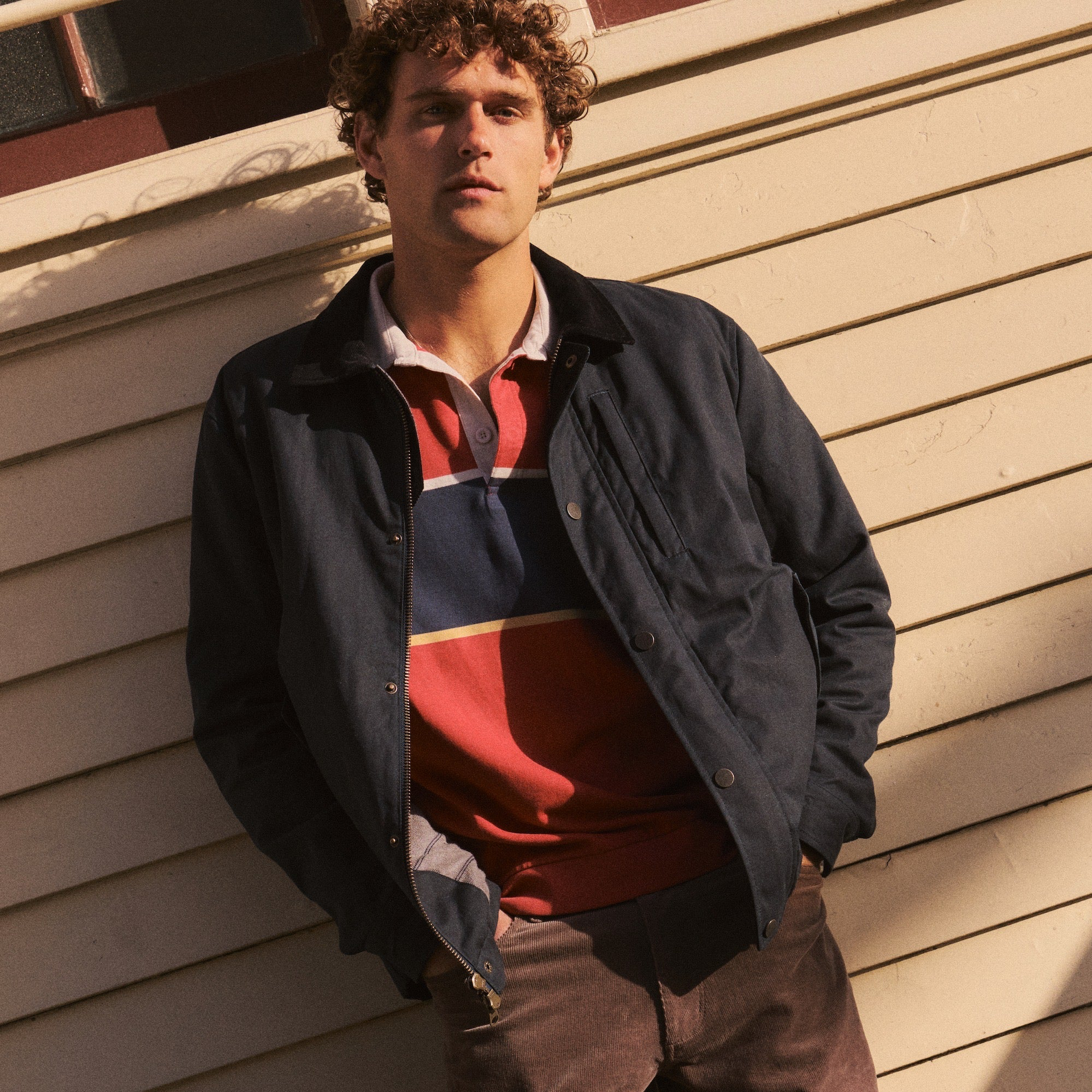 A man wearing a dark navy waxed cotton utility jacket over a red, navy, and white striped polo shirt, leaning against a beige wood-paneled wall.