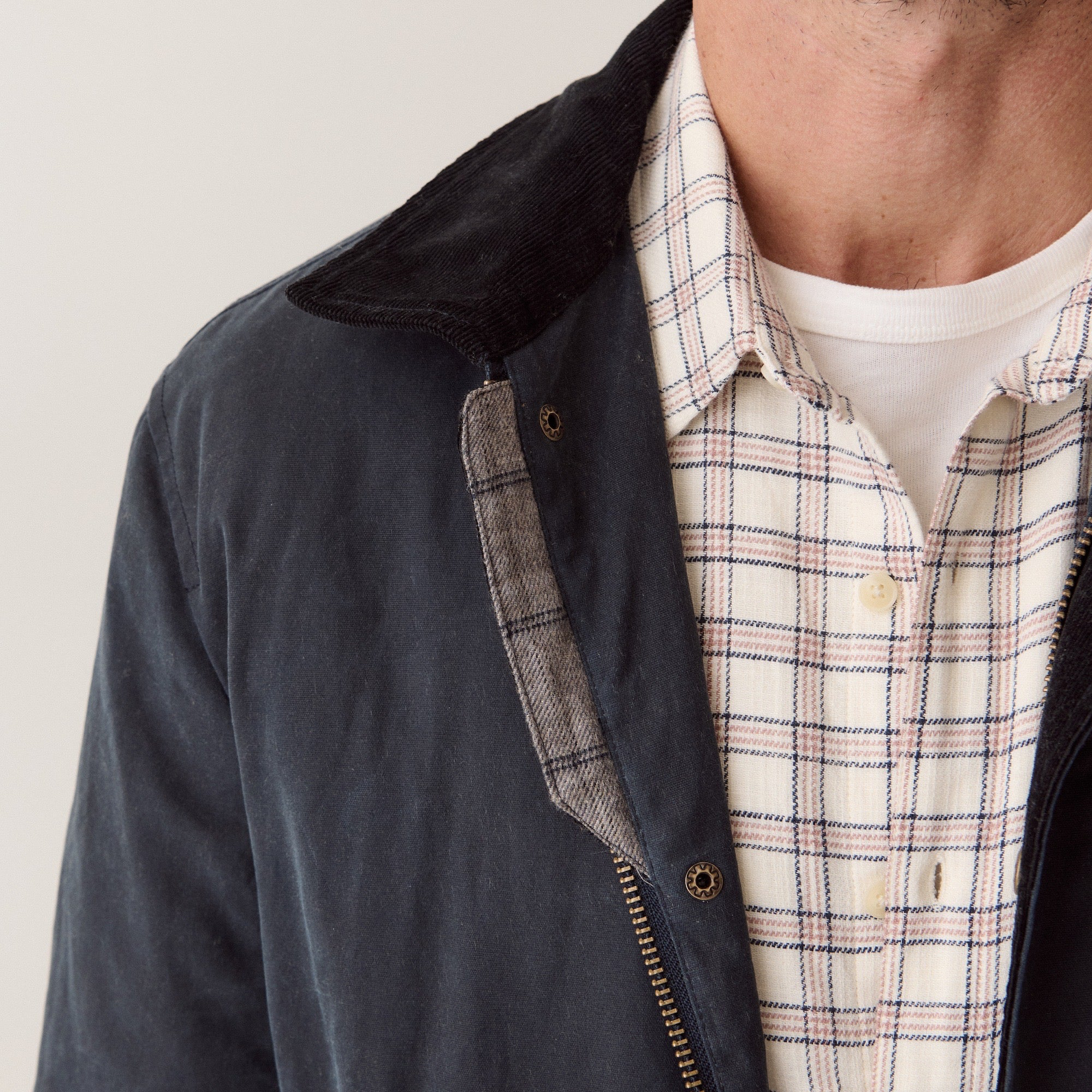 Close-up of a person wearing a dark navy jacket with a corduroy collar and zippered front, layered over a white plaid button-up shirt and a white undershirt, against a plain light background.