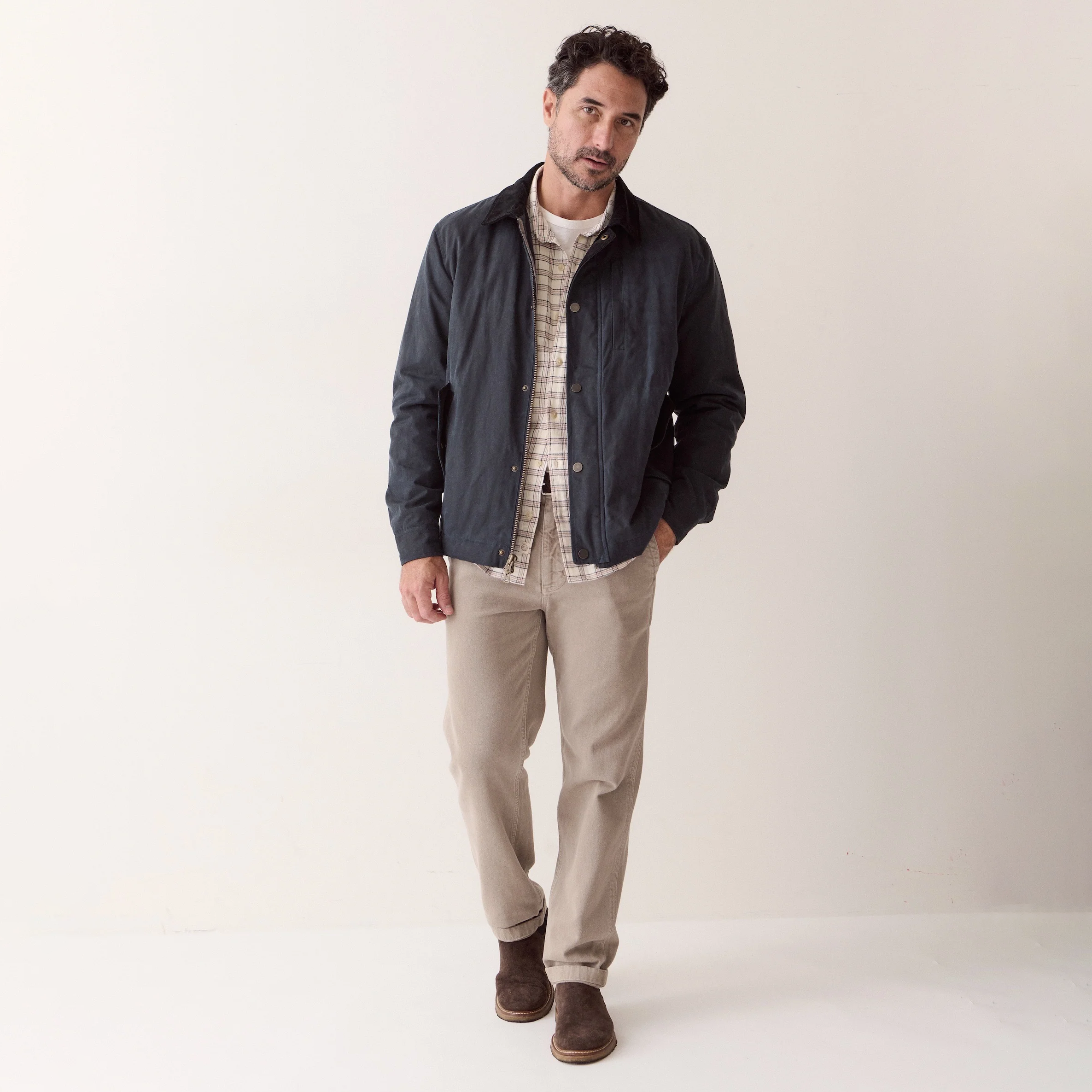A man modeling a dark navy jacket over a beige and white plaid shirt, paired with beige trousers and brown boots, standing against a plain off-white studio background.