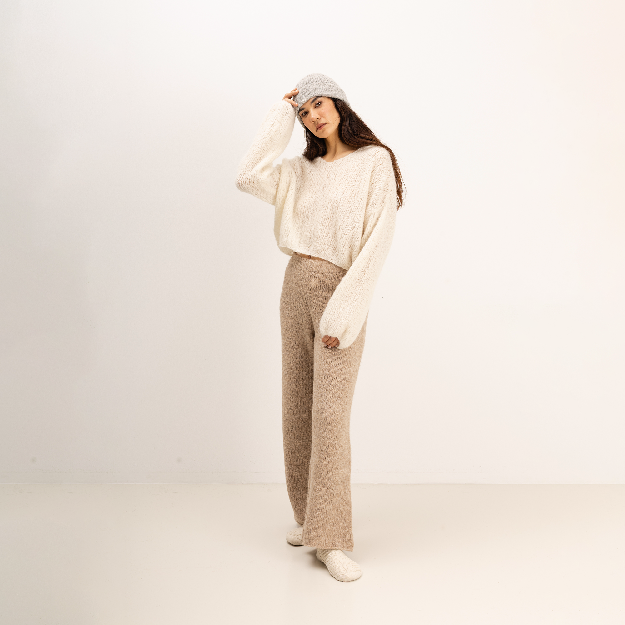 A woman modeling a cozy knit outfit consisting of a cream-colored fuzzy cropped sweater, beige wide-leg knit pants, a light gray beanie, and cream socks, set against a plain off-white studio background.