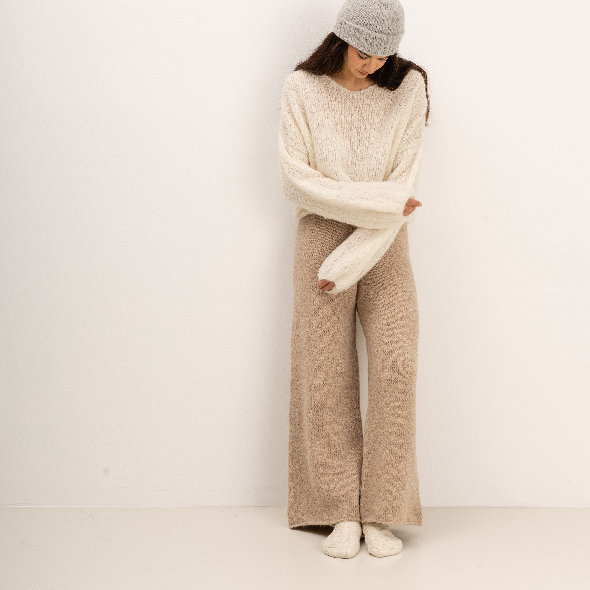 A woman stands against a plain off-white wall wearing a chunky cream knit sweater with oversized sleeves, wide-leg beige knit pants, a light gray beanie, and cream socks.