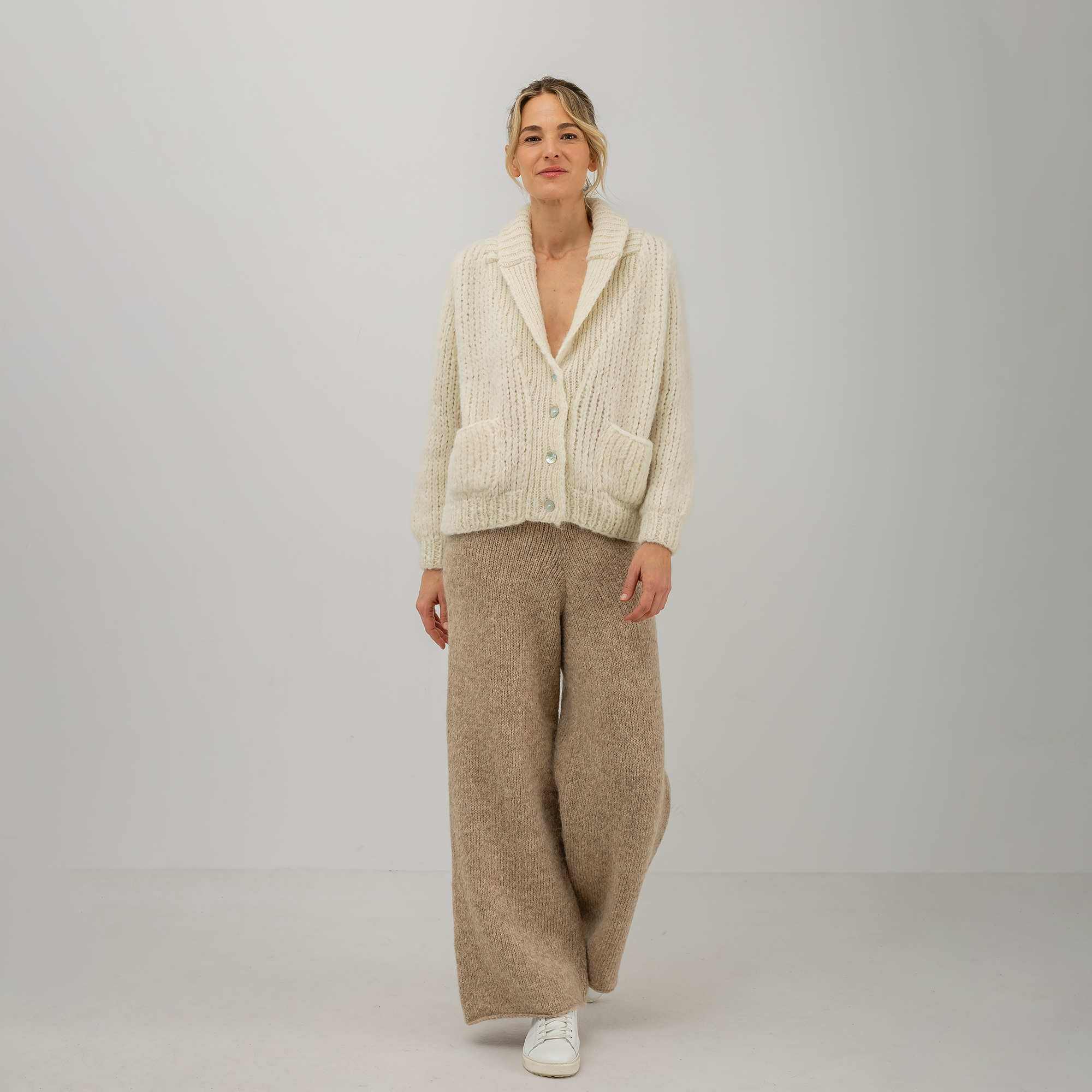 A woman modeling a cream cable-knit cardigan and beige wide-leg knit pants against a light gray studio background.