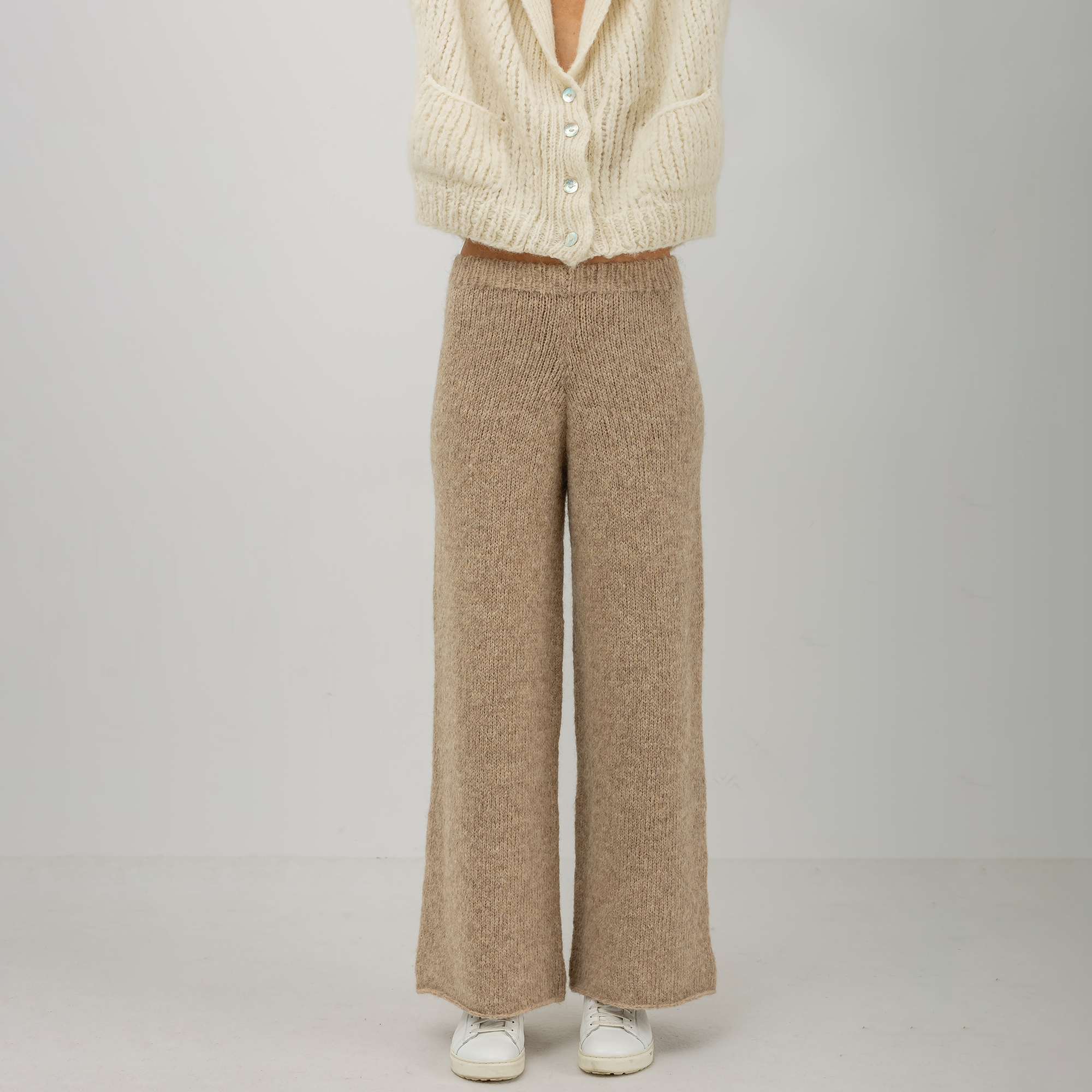 A person wearing textured knit wide-leg pants in a warm taupe-beige color, paired with a cream cardigan and white sneakers, standing against a light gray studio background.