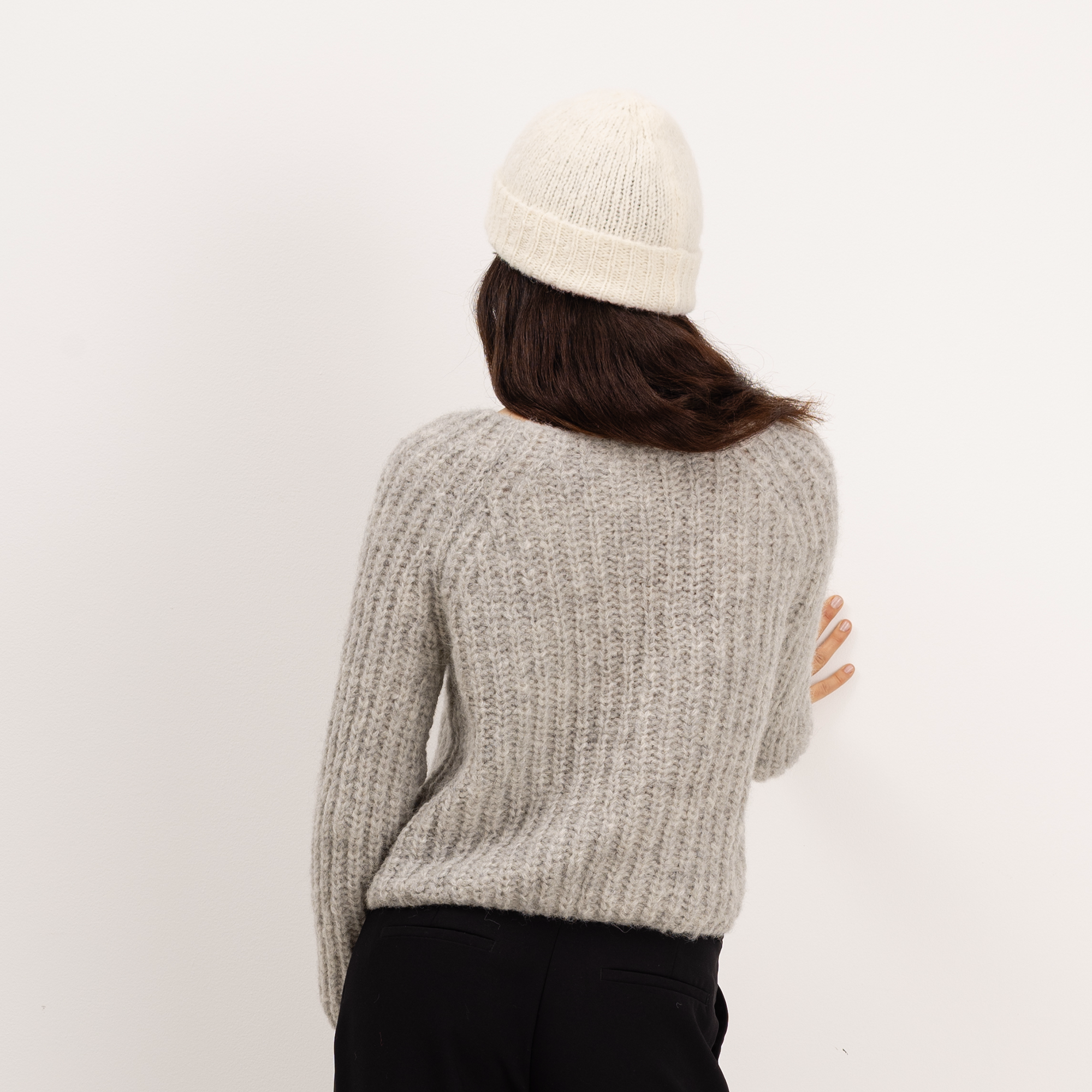 A person shown from the back wearing a light gray ribbed knit sweater, a cream beanie, and black pants against a plain white background.