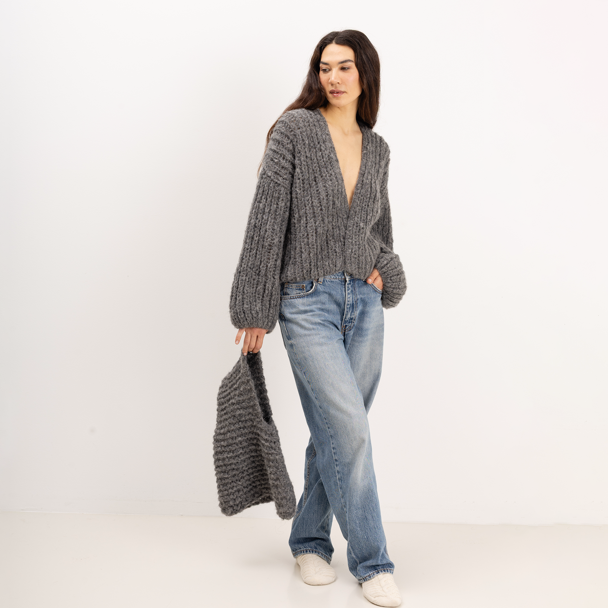 A woman stands against a plain white studio background wearing a chunky gray knit cardigan with a deep V-neck and long balloon sleeves, paired with light blue jeans and white slip-on shoes. She is holding a matching gray knit bag.