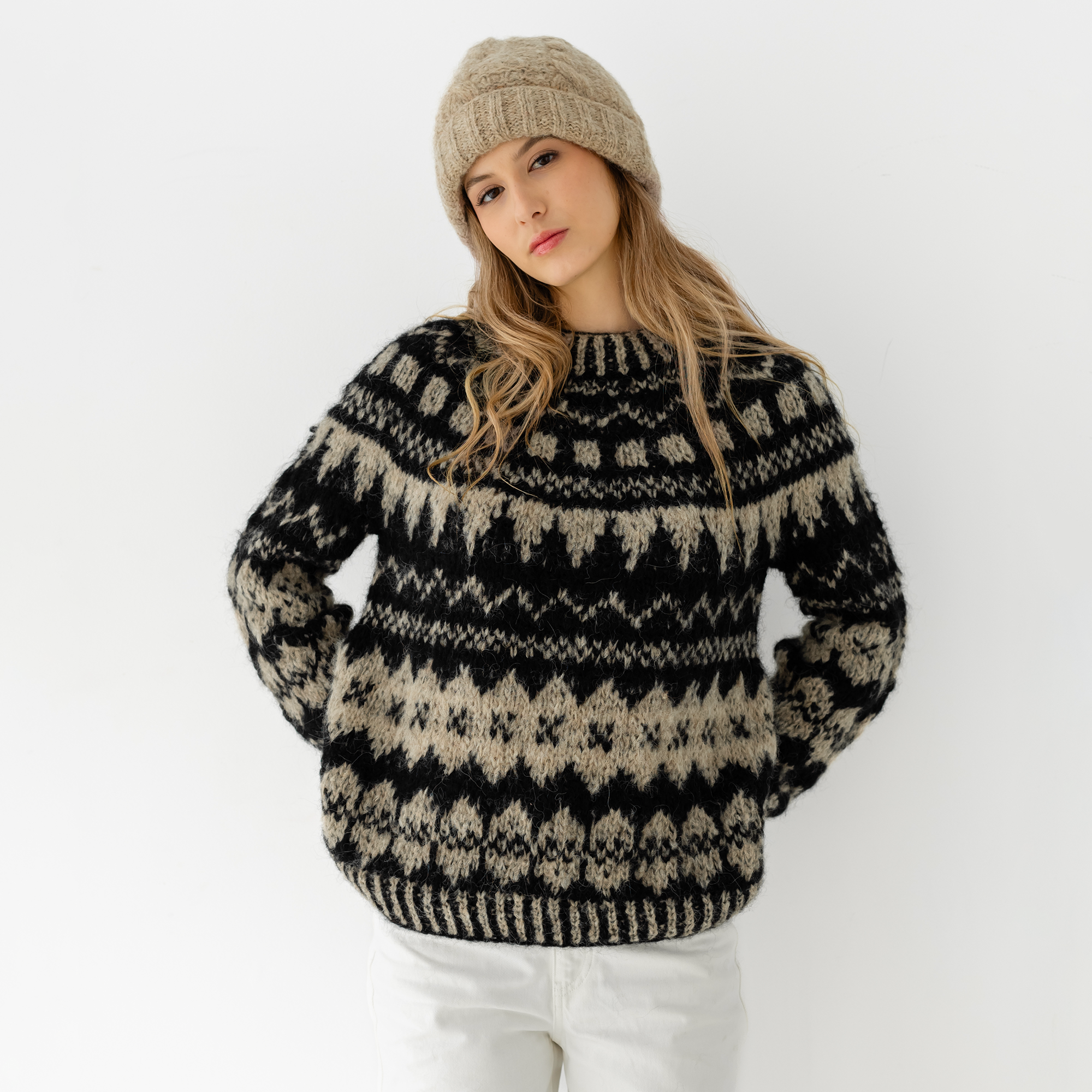 A woman wearing a black and beige patterned knit sweater with a matching beige knit beanie, posed against a plain white studio background.