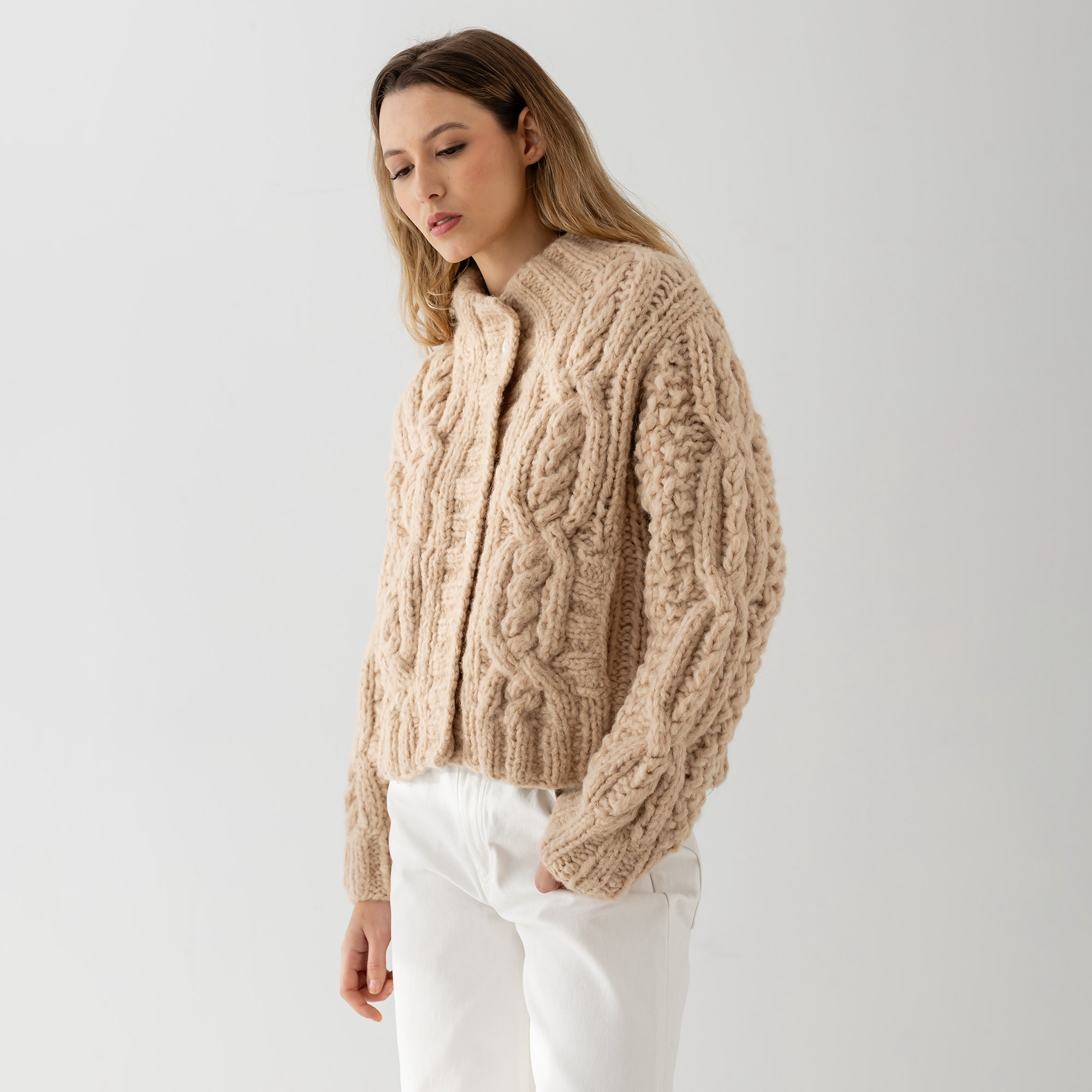 A woman wearing a beige cable-knit cropped cardigan with white pants, photographed against a plain light gray studio background.