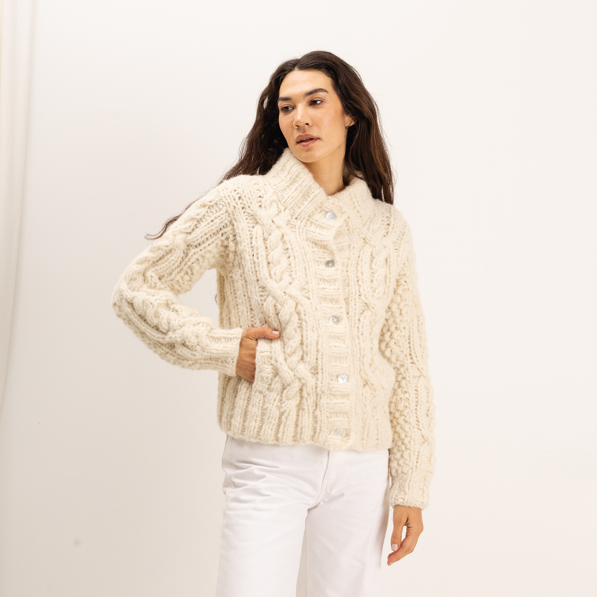 A woman wearing a cream-colored, chunky cable-knit cardigan sweater with white pants, standing against a plain white studio background.