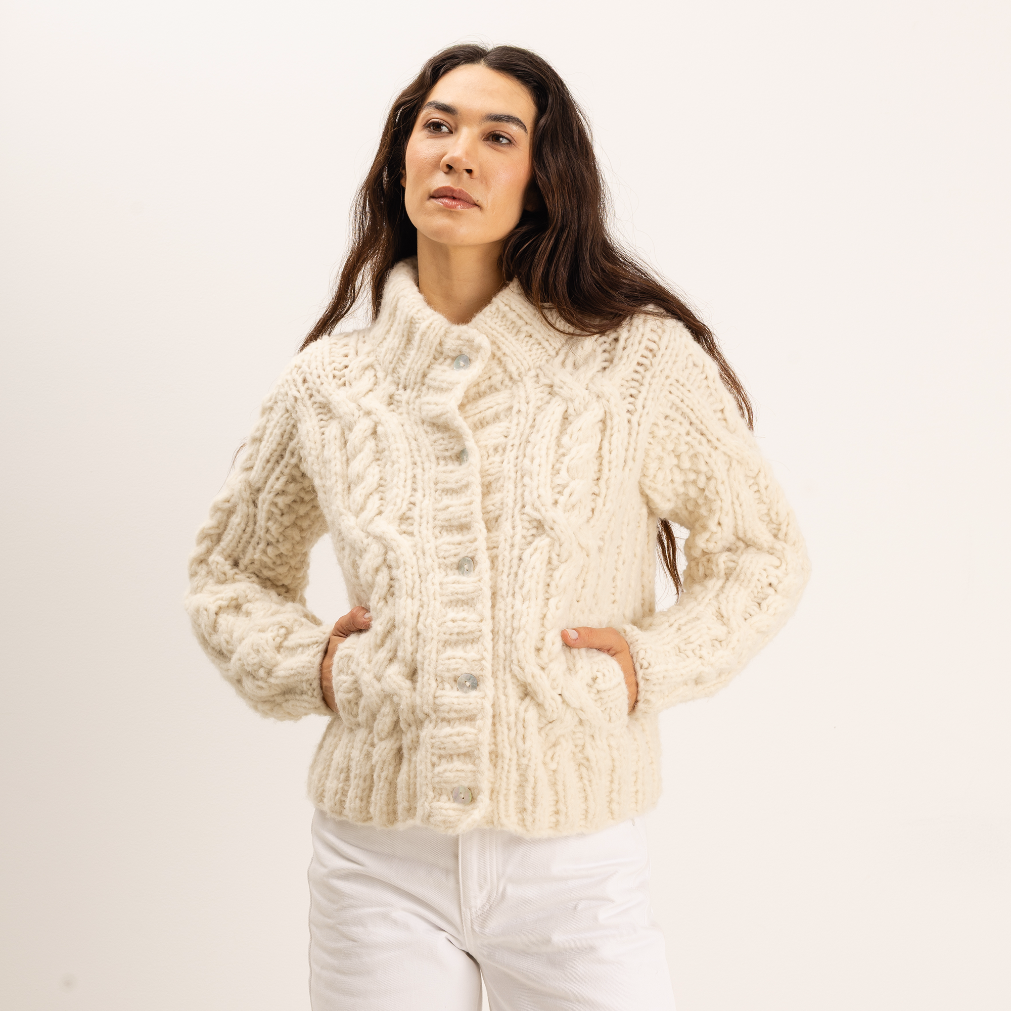 A woman wearing a chunky cream cable-knit cardigan sweater with white pants, standing against a plain off-white studio background.