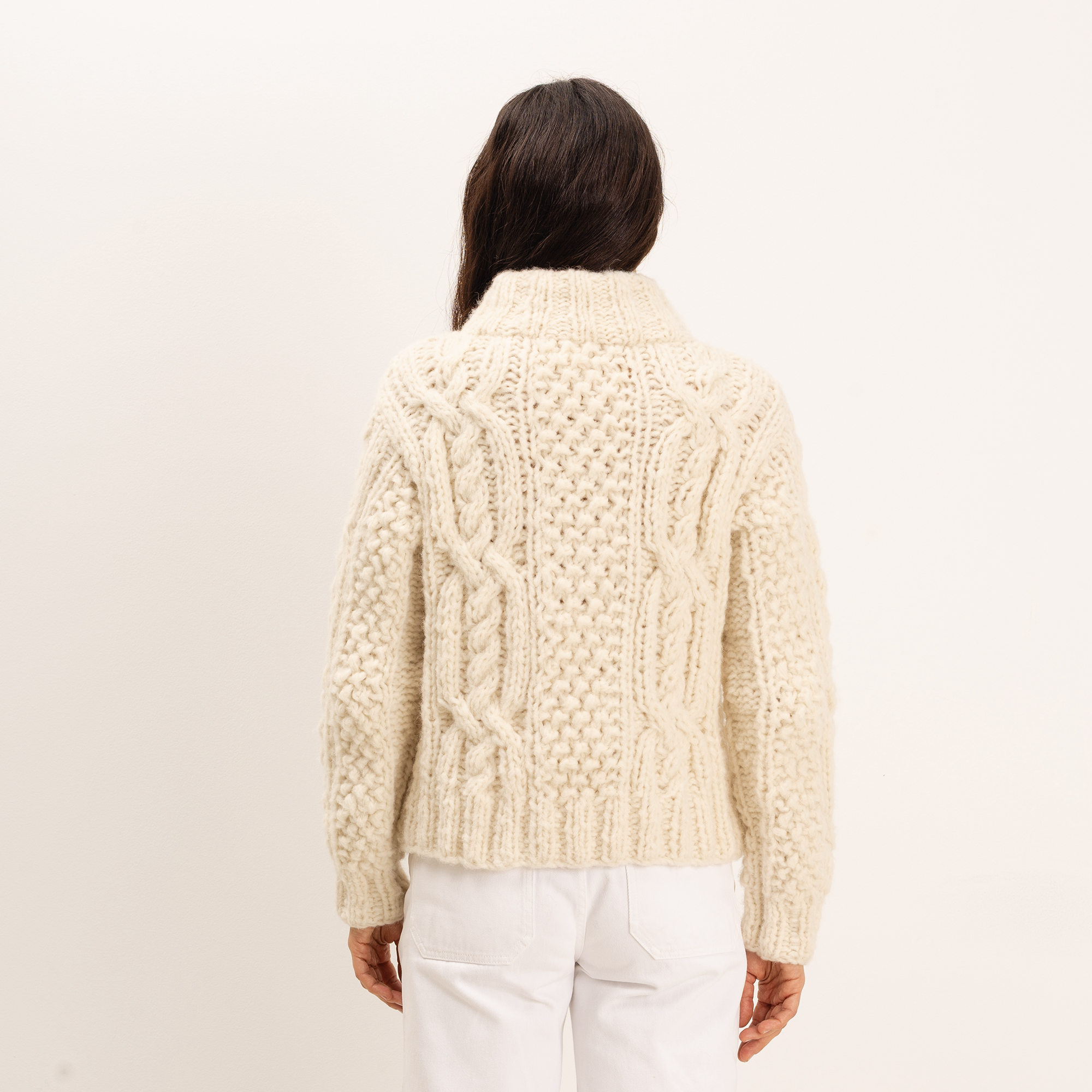 A woman viewed from behind wearing a cream cable-knit cropped sweater and white pants against a plain off-white studio background.