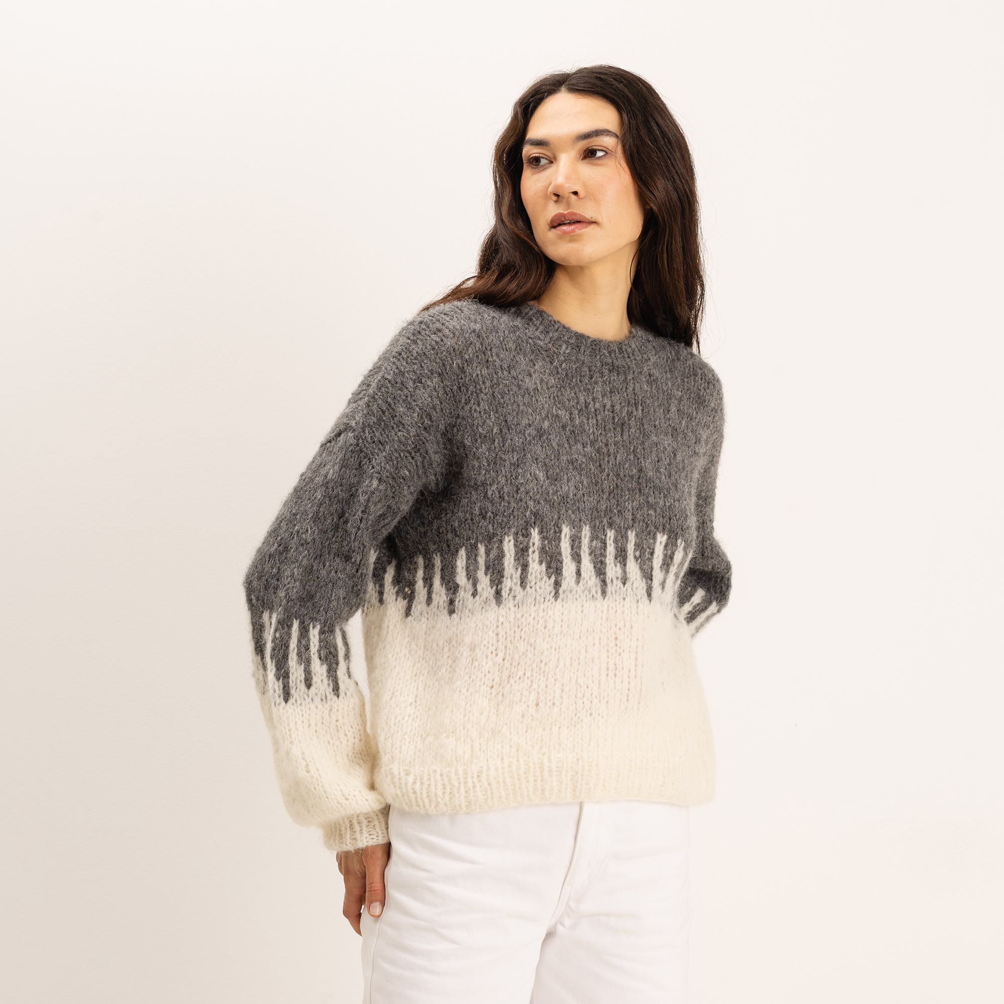 A woman modeling a fuzzy knit sweater with a dark gray upper section and a cream lower section with a vertical drip-like pattern, paired with white pants against a plain off-white background.