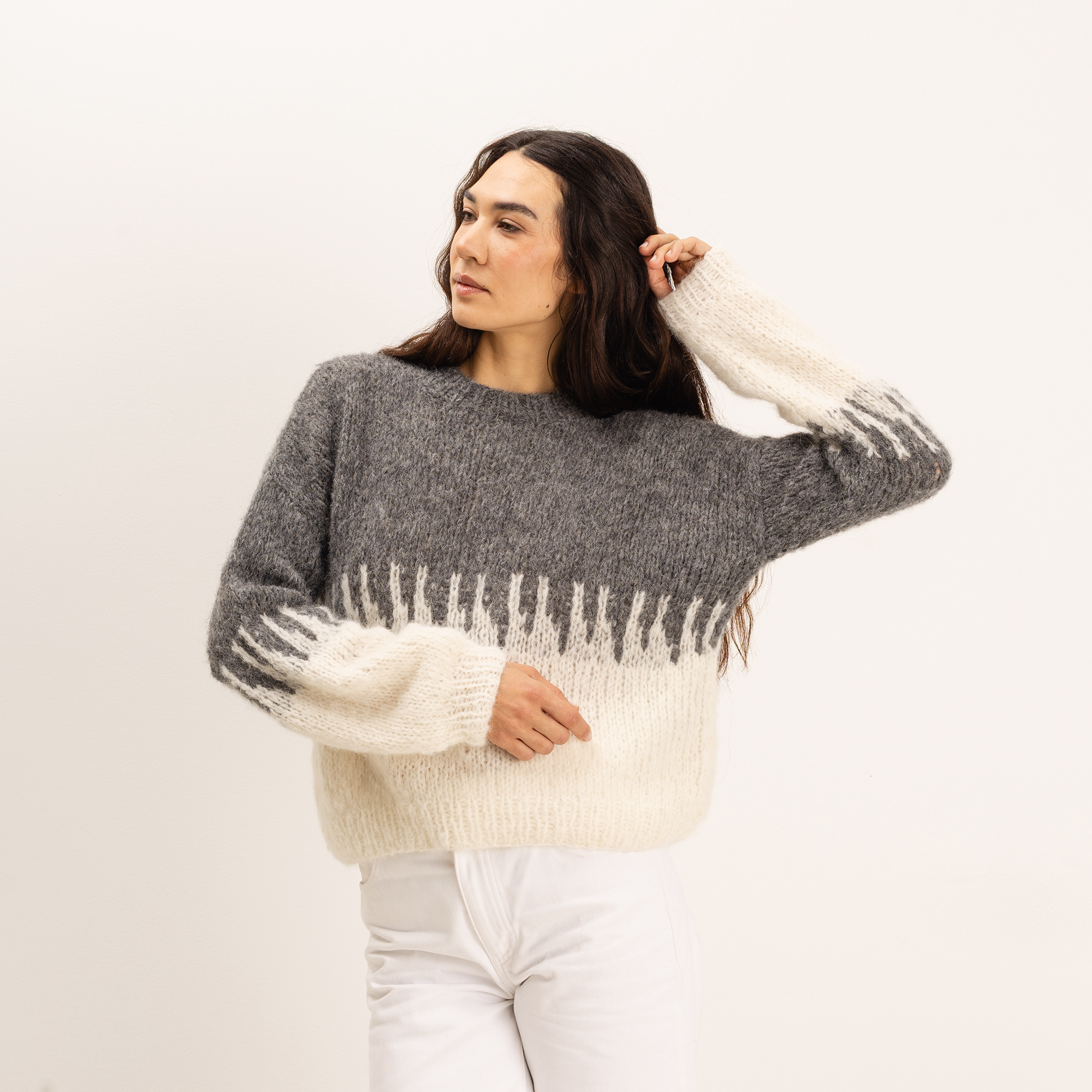 A woman modeling a fuzzy knit sweater with a gray upper section and an off-white lower section and cuffs, paired with white pants against a plain light background.