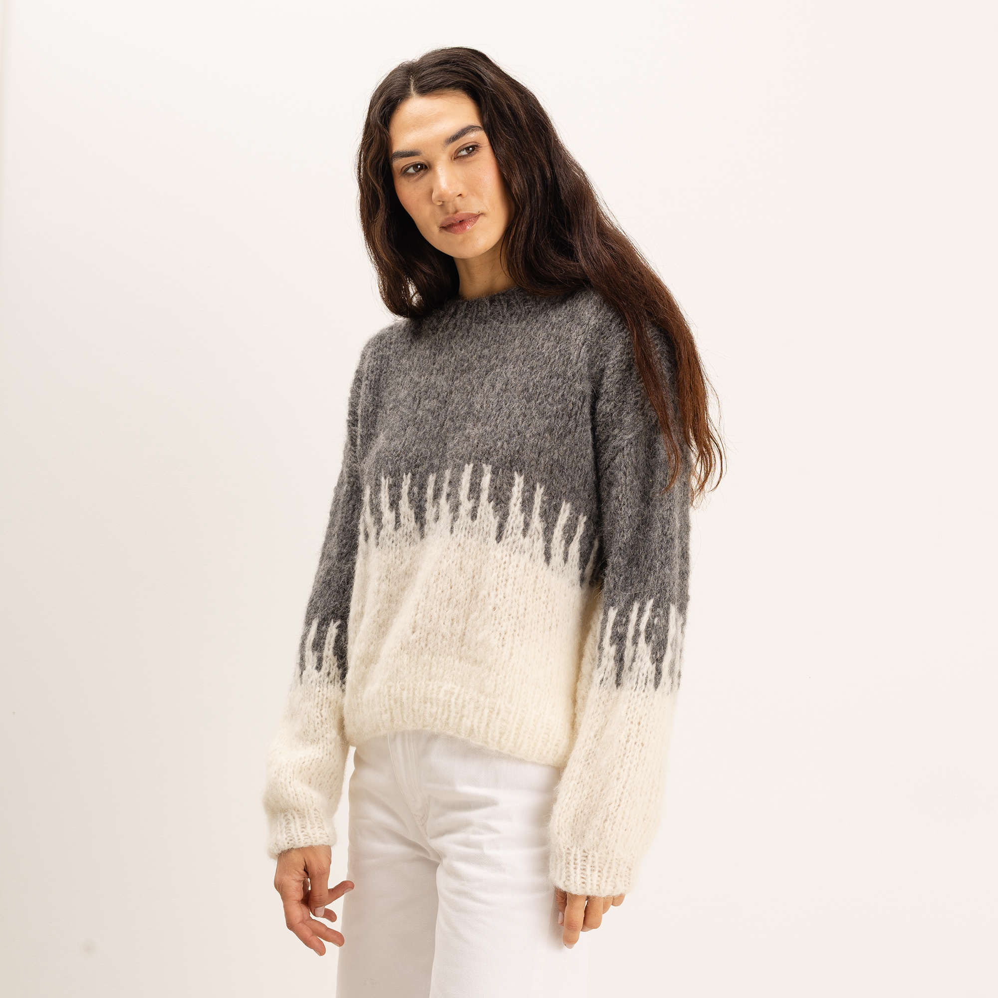 A woman modeling a fuzzy knit sweater with a color-blocked design in dark gray and cream, paired with white pants, against a plain white studio background.