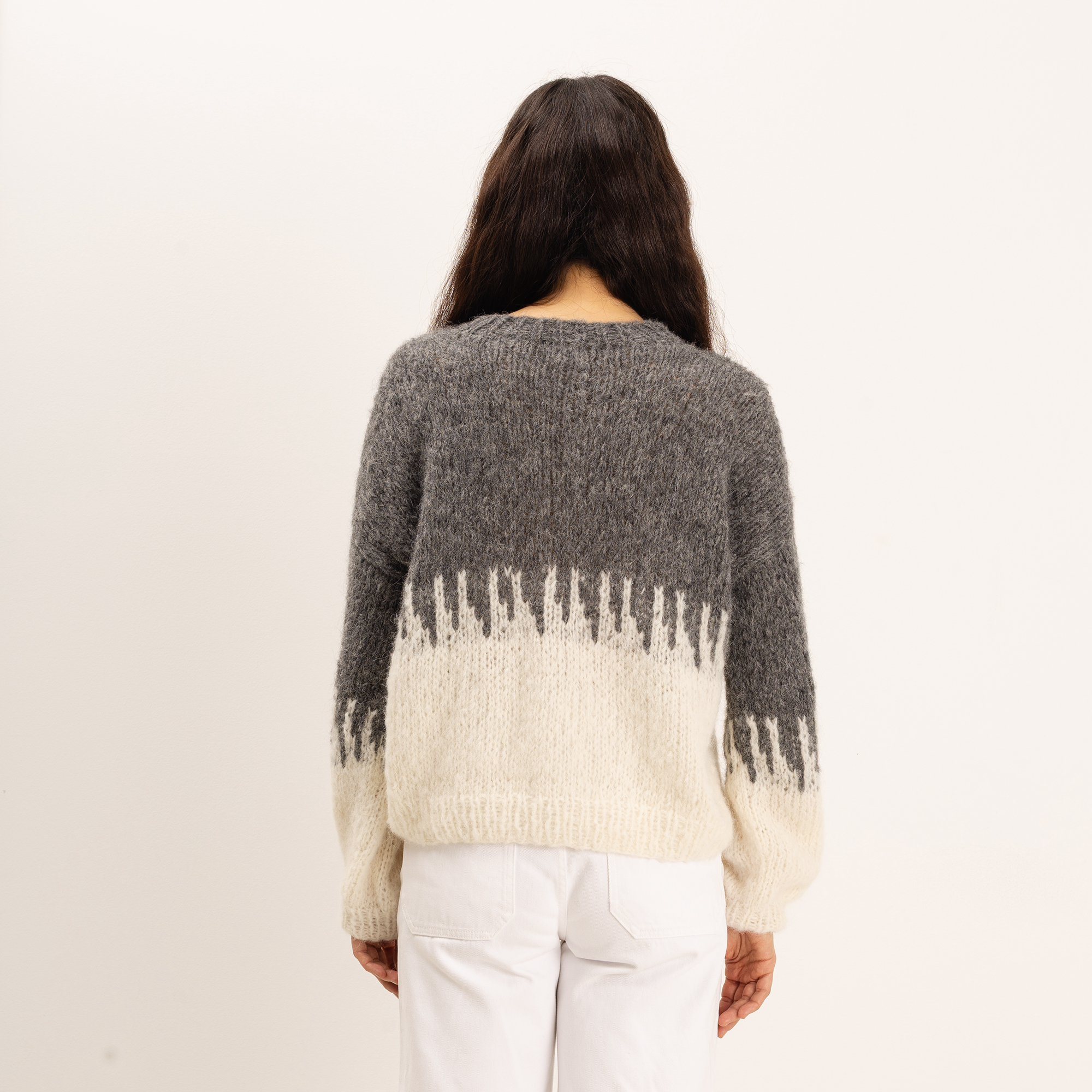 A woman viewed from behind wearing a fuzzy knit sweater with a dark gray upper section and a cream lower section with a jagged horizontal pattern, paired with white pants against a plain off-white background.