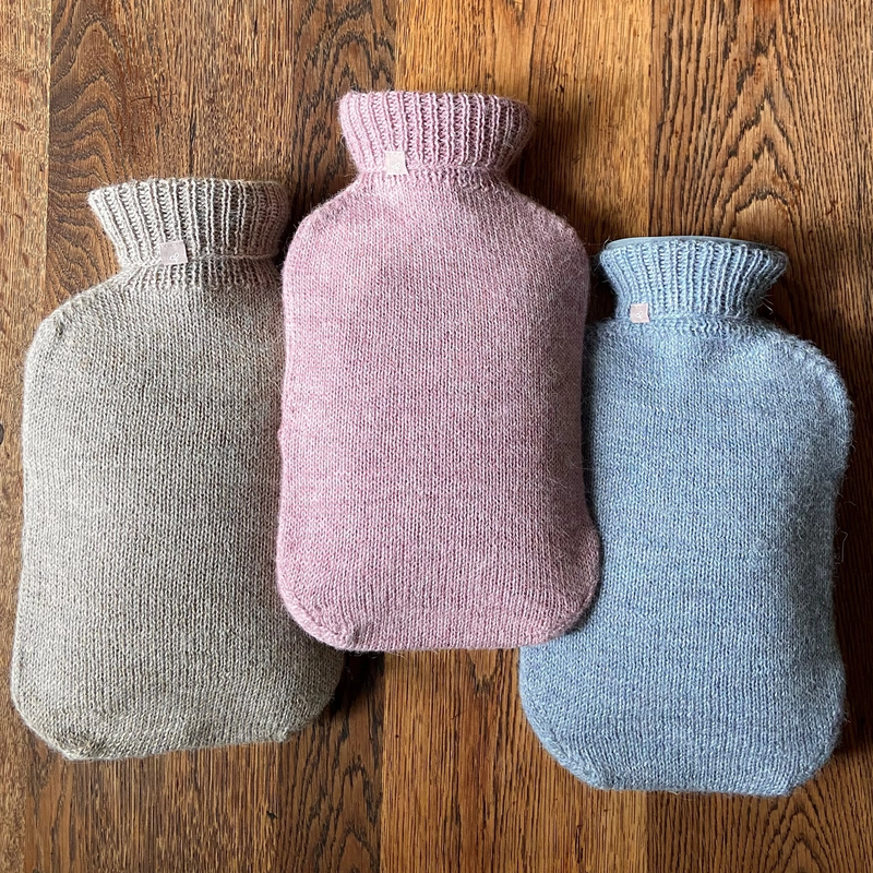 Three alpaca wool hot water bottle covers in beige, pink, and blue colors, displayed on a wooden floor.