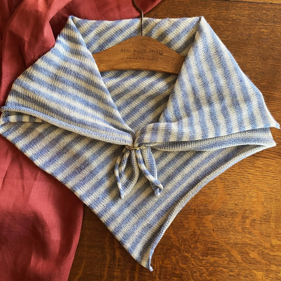 A striped alpaca wool neckerchief in blue and white tones, displayed on a wooden surface with a red garment partially visible on the left side.