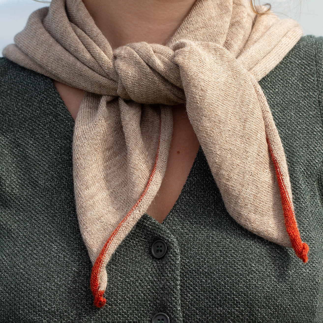 A close-up of a person wearing a light beige scarf tied around the neck, with bright orange-red striped ends and a dark green knit cardigan.