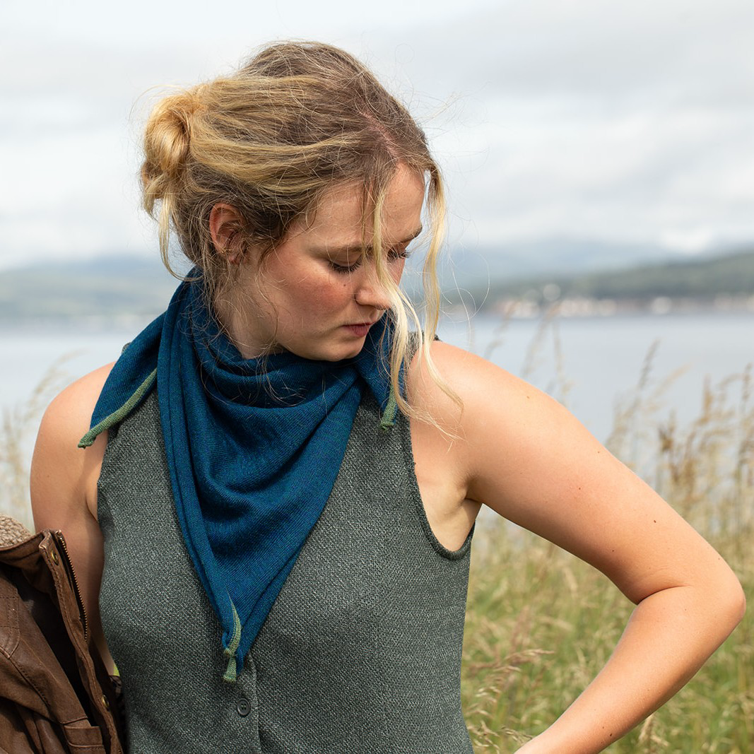A woman outdoors by the water wearing a dark teal blue triangular scarf wrapped around her neck, paired with a gray sleeveless top.