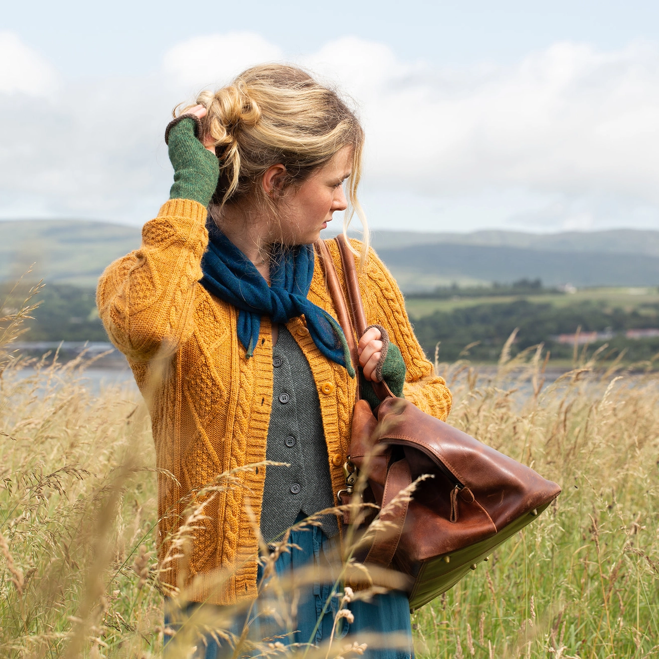 A woman standing in a grassy field outdoors, wearing a chunky cable-knit mustard yellow cardigan, a teal scarf, green knit gloves, and carrying a brown leather shoulder bag.