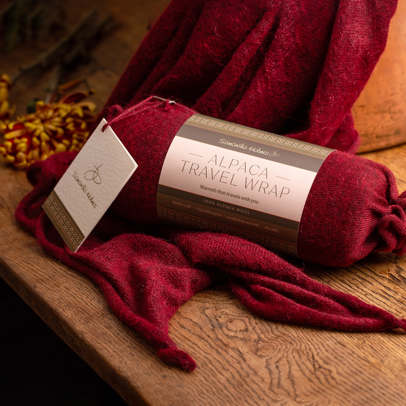 A rolled Alpaca Travel Wrap scarf with a paper label and hanging tag, resting on a rustic wooden surface with a draped maroon wrap around it.