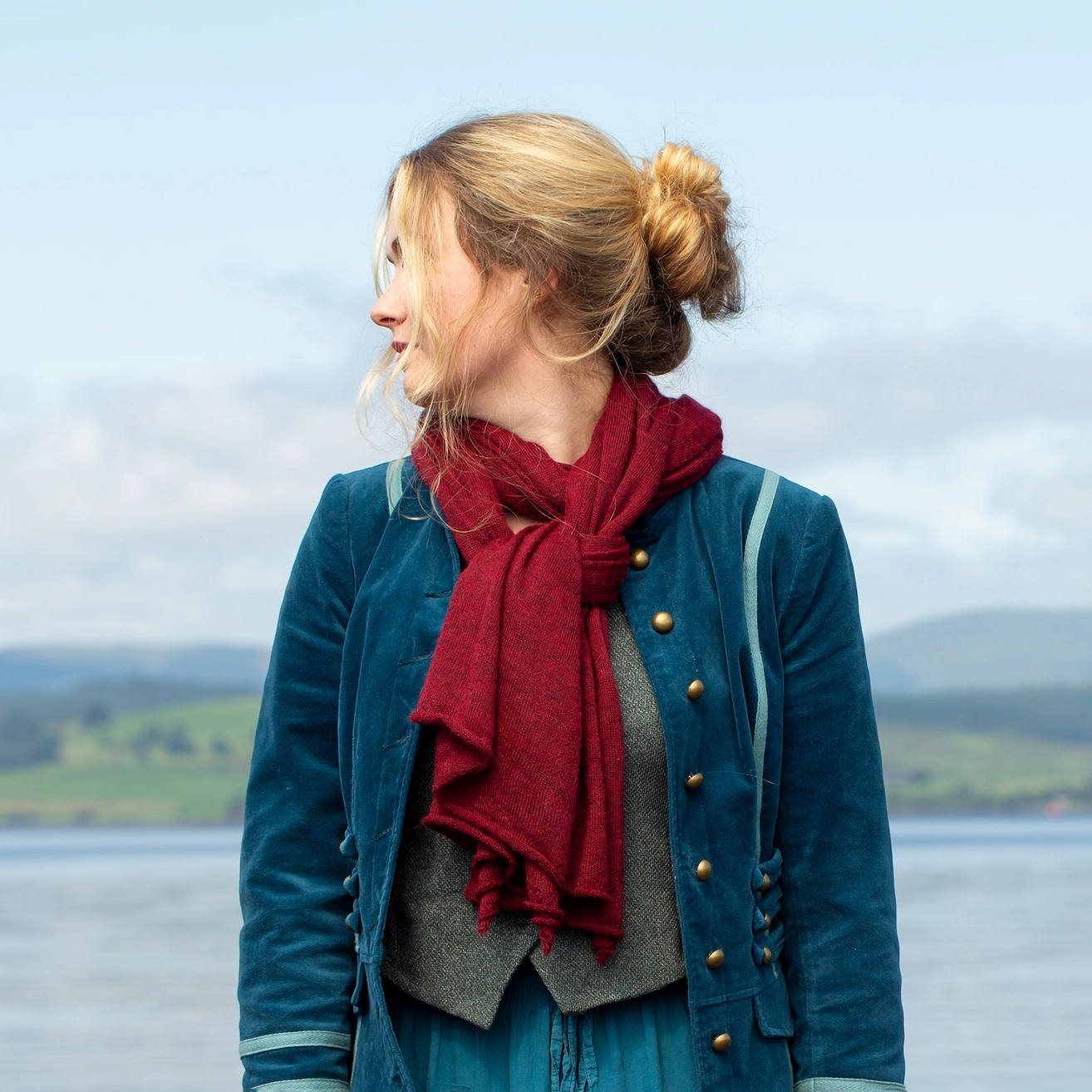 A woman in profile wearing a teal blue velvet jacket, gray vest, and a deep red scarf, standing outdoors with a blurred lake and hills in the background.