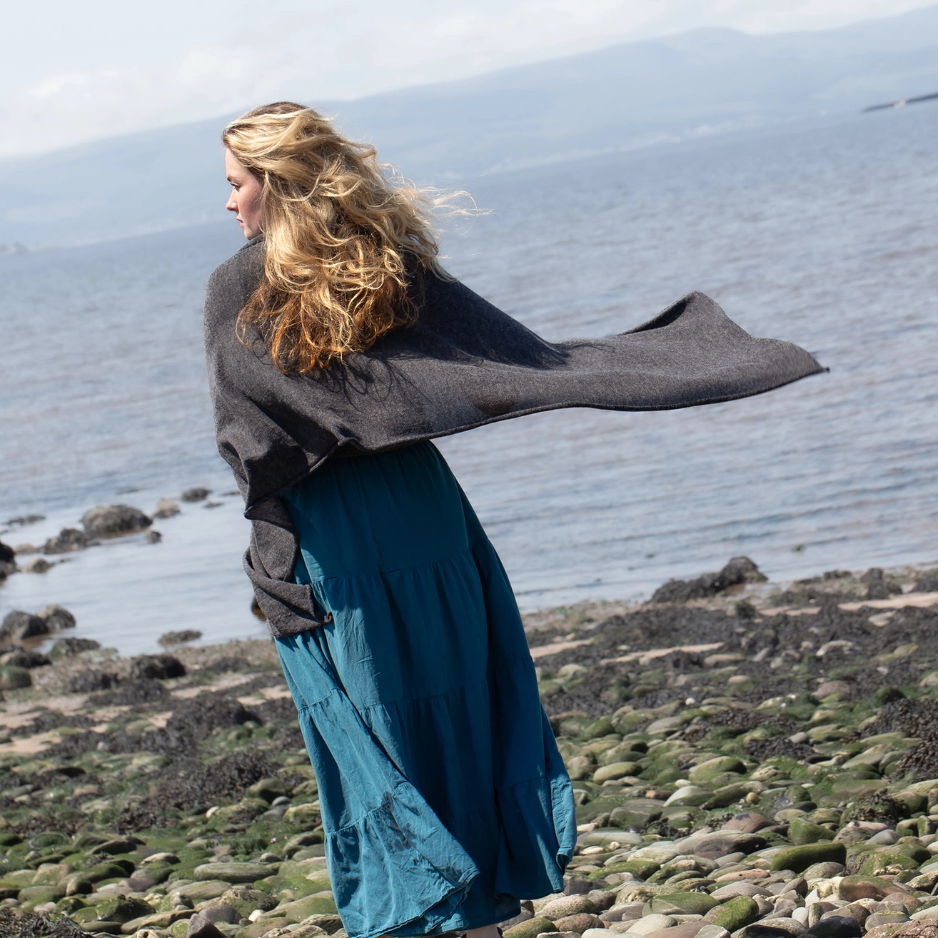 A woman stands on a rocky shoreline with the sea and distant mountains in the background, wearing a dark charcoal gray shawl draped over her shoulders and a long teal-blue skirt.