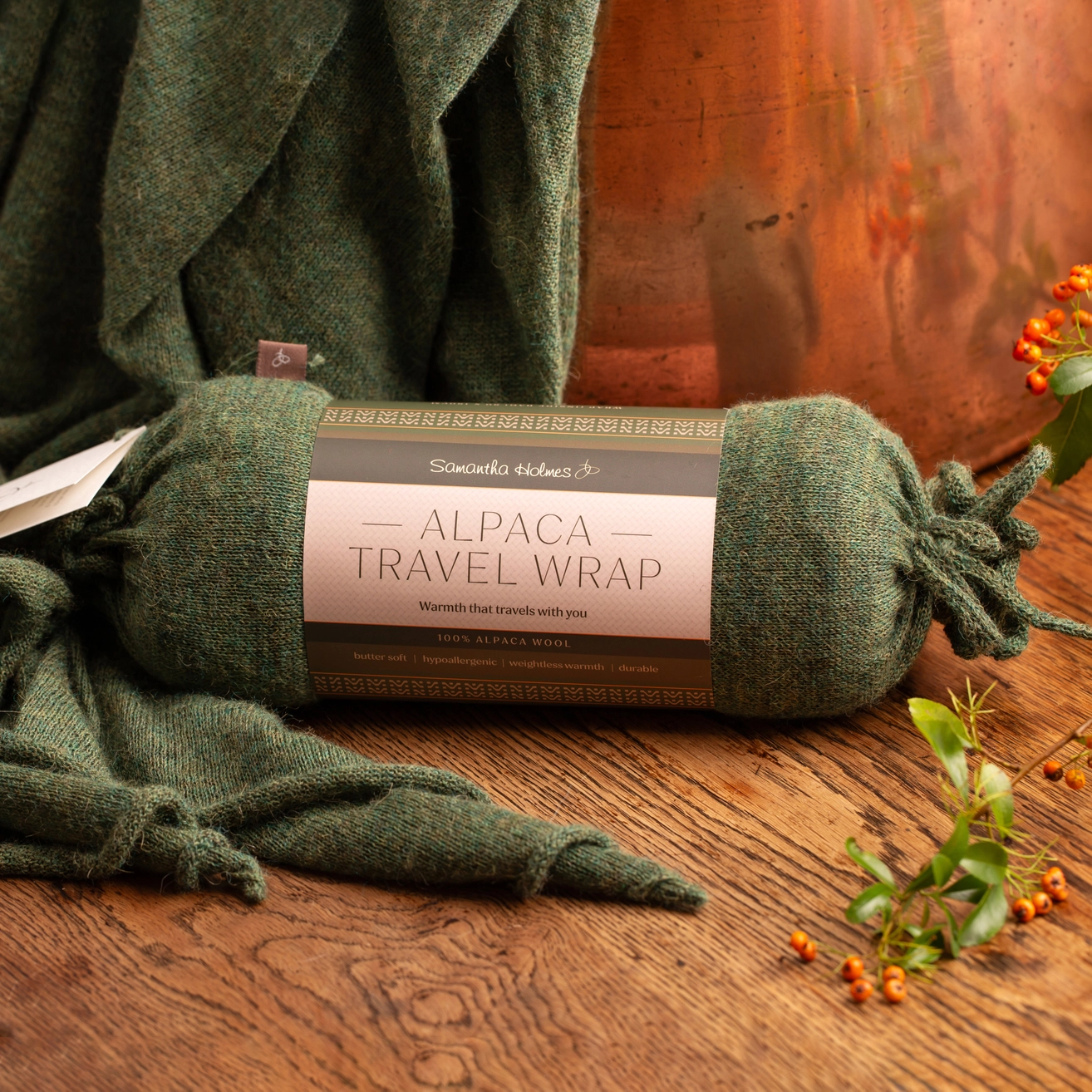 A green Alpaca Travel Wrap styled on a rustic wooden table with a copper pot in the background and small orange berries in the foreground.