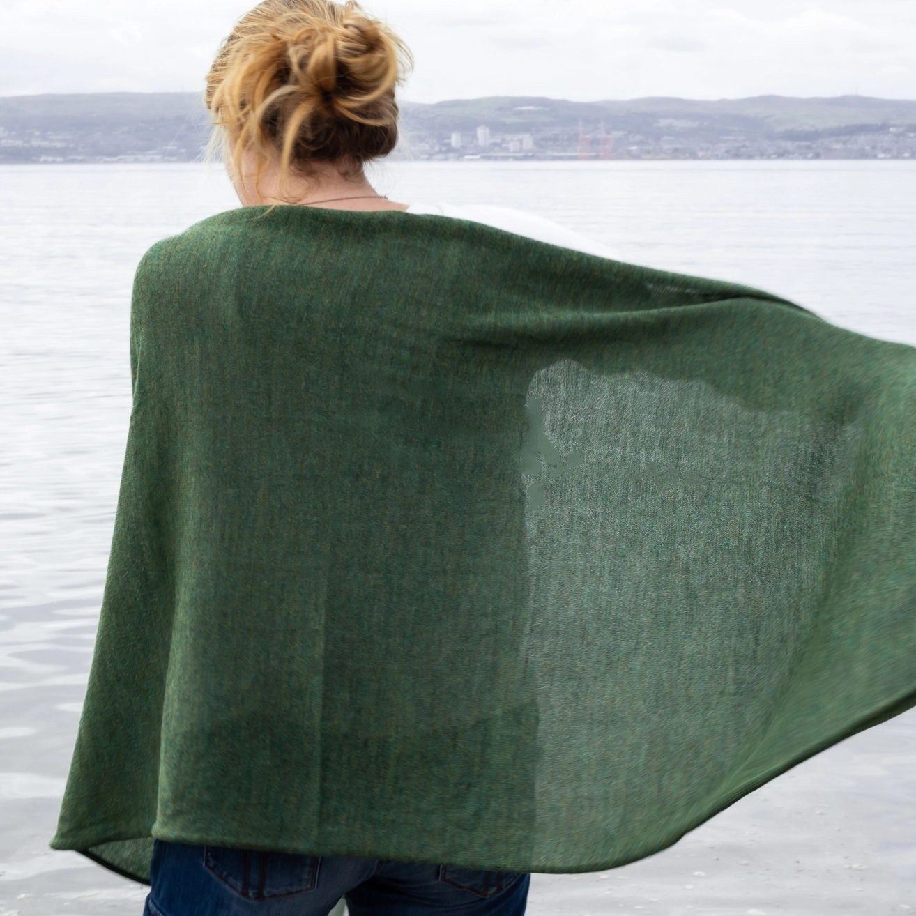 A woman seen from behind wearing a flowing dark green shawl outdoors by the water, with a blurred shoreline in the background.