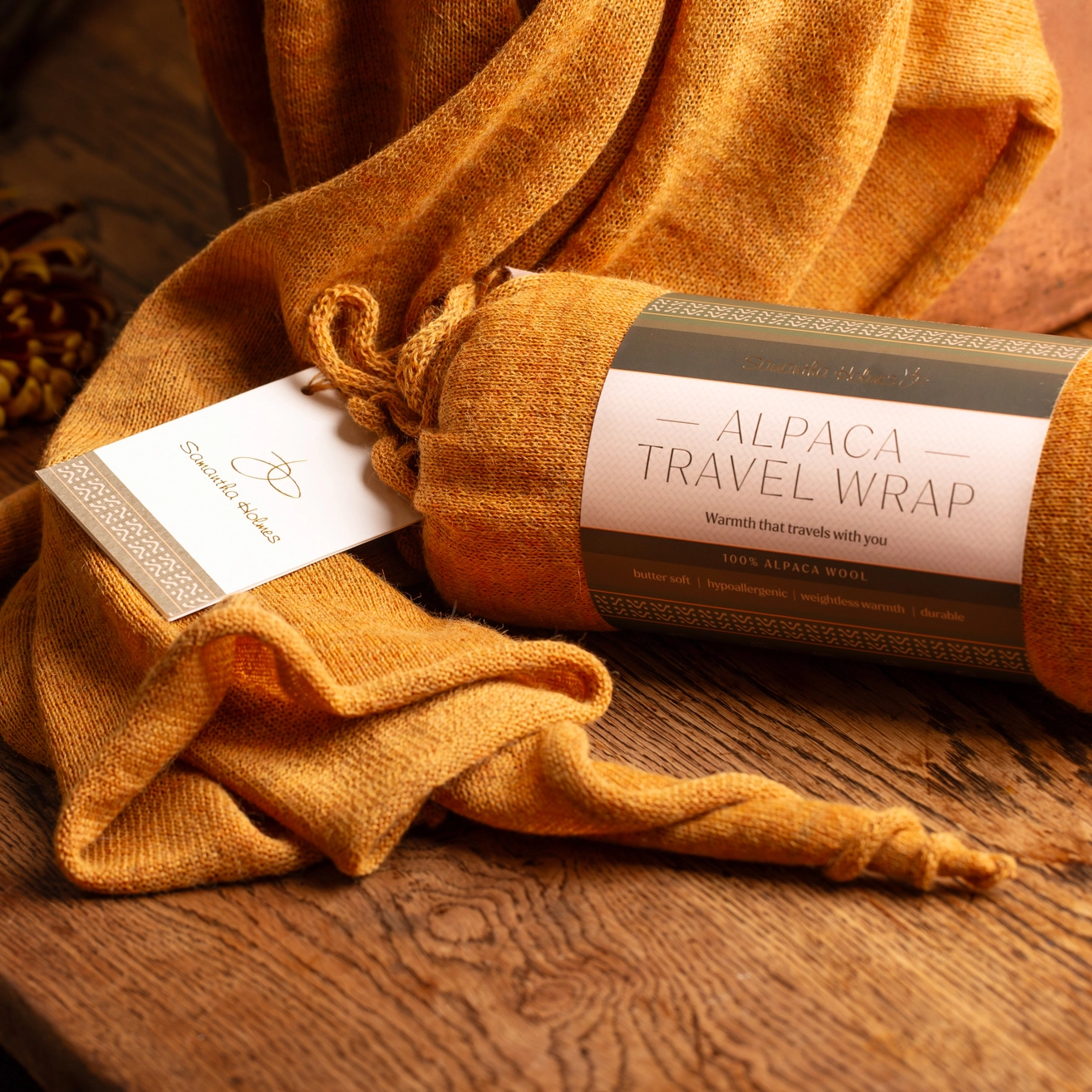 A rolled and draped mustard-yellow alpaca travel wrap with a paper tag and wrapped label, styled on a rustic wooden surface with dried botanicals in the background.