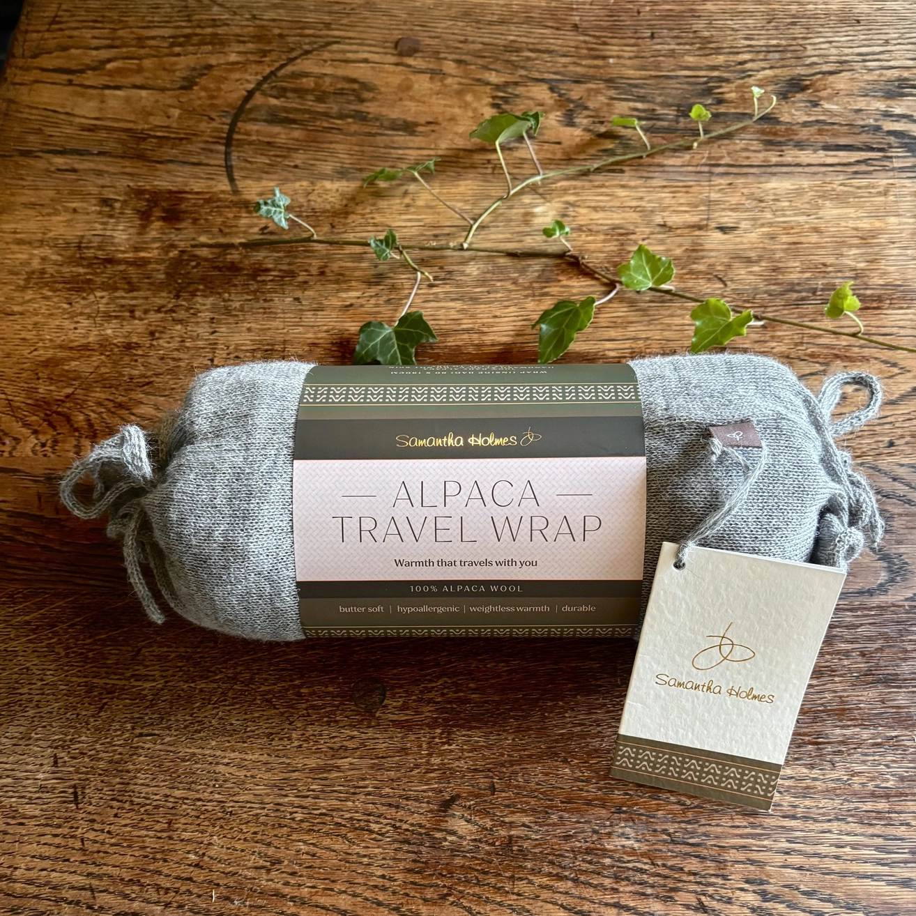 A rolled gray Alpaca Travel Wrap displayed on a rustic wooden table with green ivy vines. The product has a dark brown paper label wrapped around it and a small paper hangtag attached.