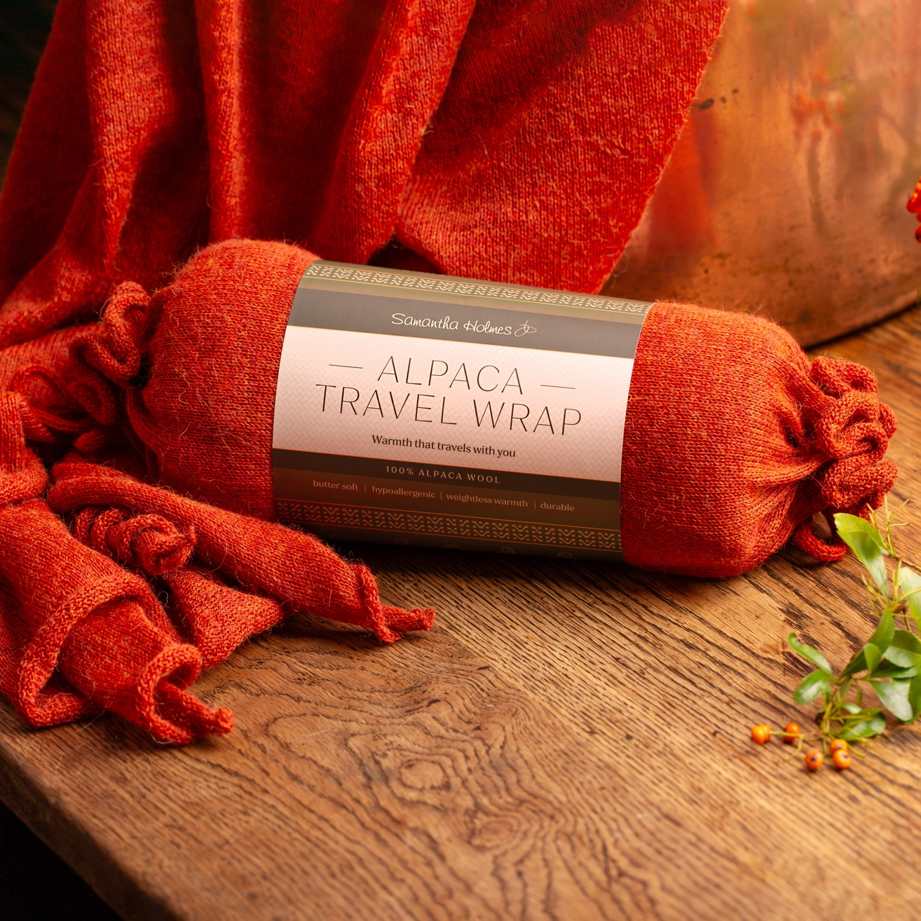 A rolled orange-red alpaca travel wrap with a paper label lies on a wooden surface, with matching fabric draped around it and a copper pot in the background.