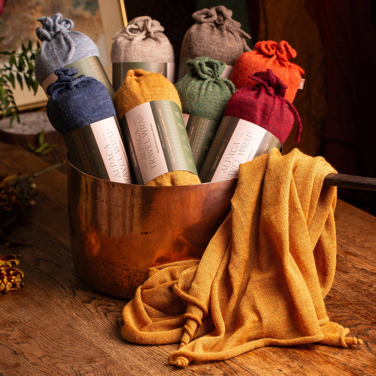 A group of colorful travel wraps and a draped yellow wrap arranged in a copper pot on a rustic wooden table. The shawls appear in muted blue, taupe, mustard yellow, sage green, burgundy, and rust orange.