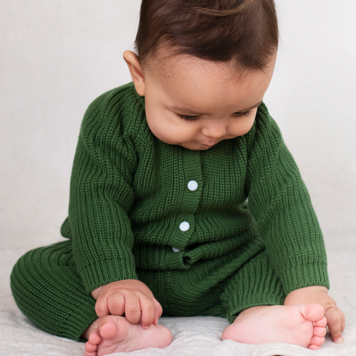 A baby wearing a green ribbed knit cardigan and matching pants, sitting on a light gray blanket against a neutral light gray background.