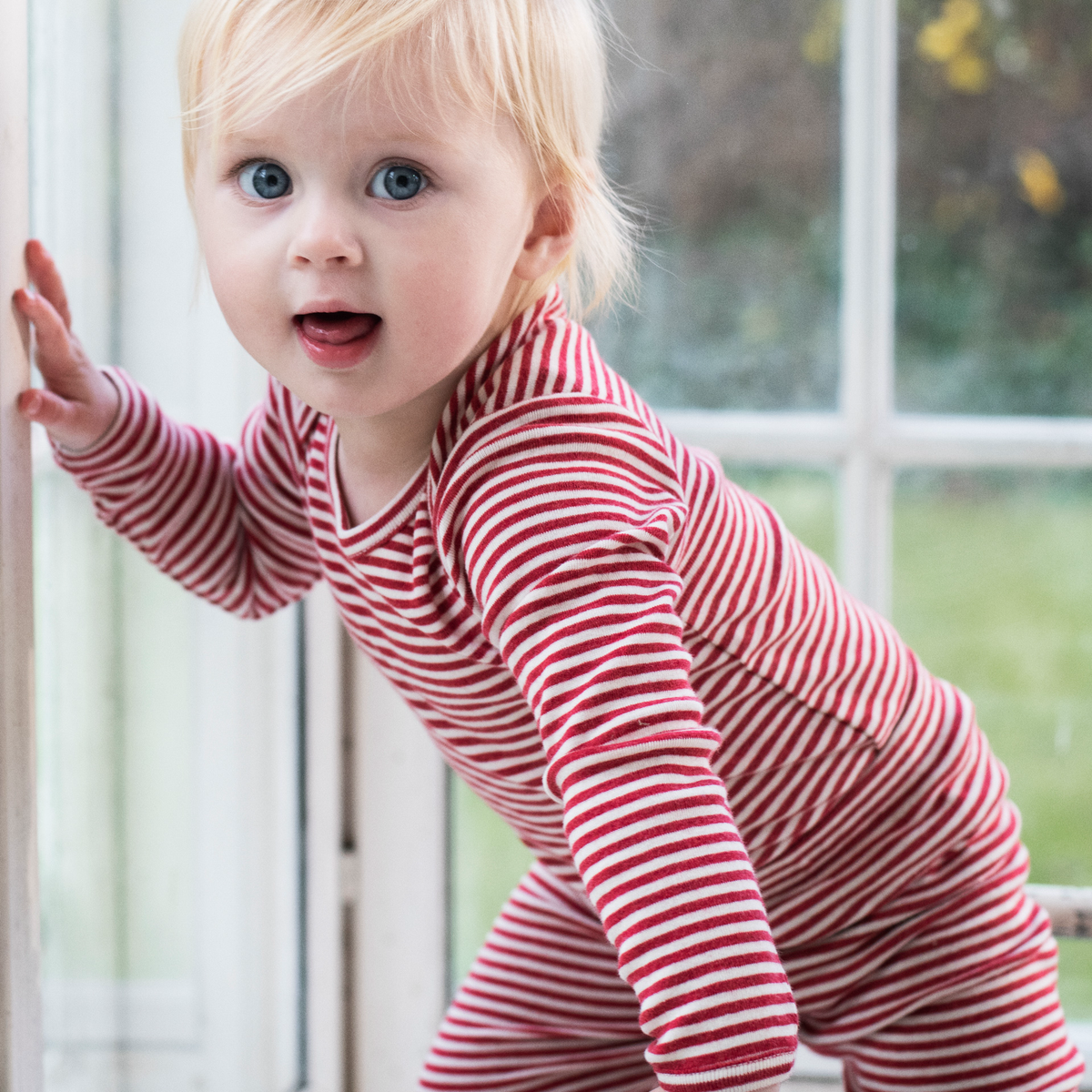 A baby toddler wearing a red and white striped long-sleeve pajama top and matching pants, leaning against a window frame in a bright indoor setting.