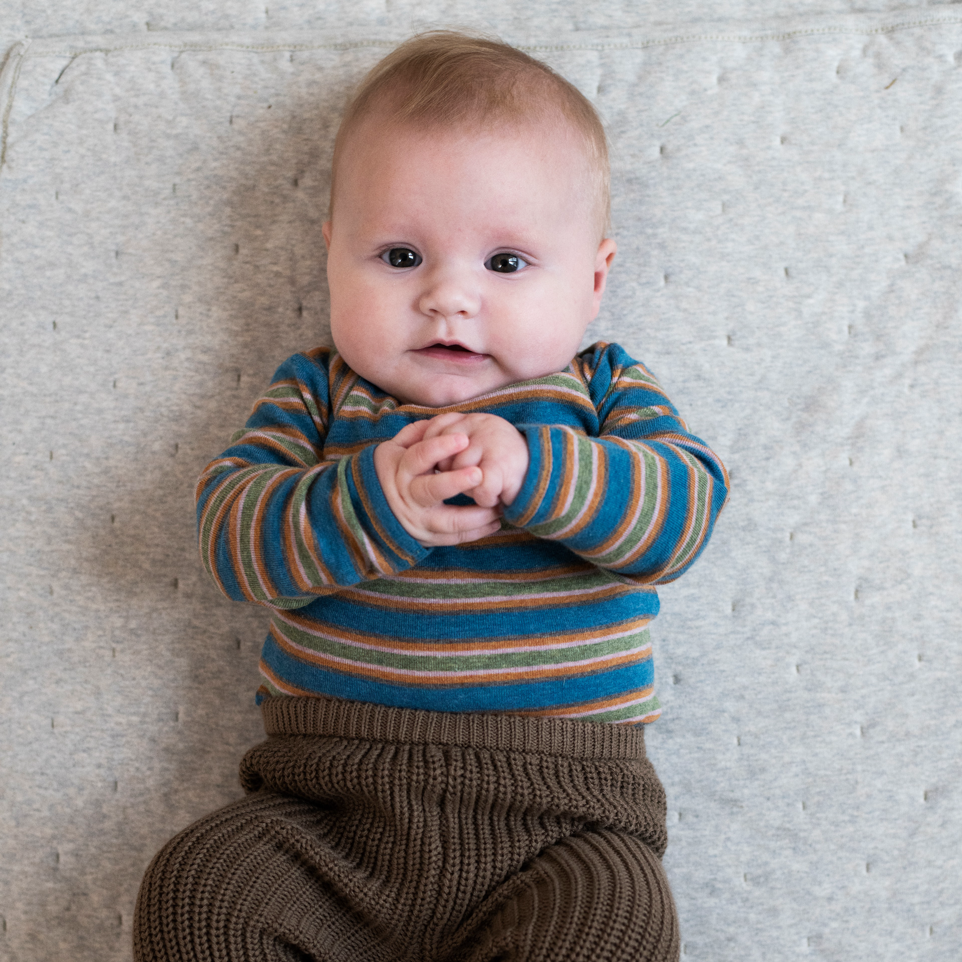 A baby lying on a textured light gray blanket, wearing a long-sleeve striped knit top in teal, brown, and beige, paired with brown knit pants.