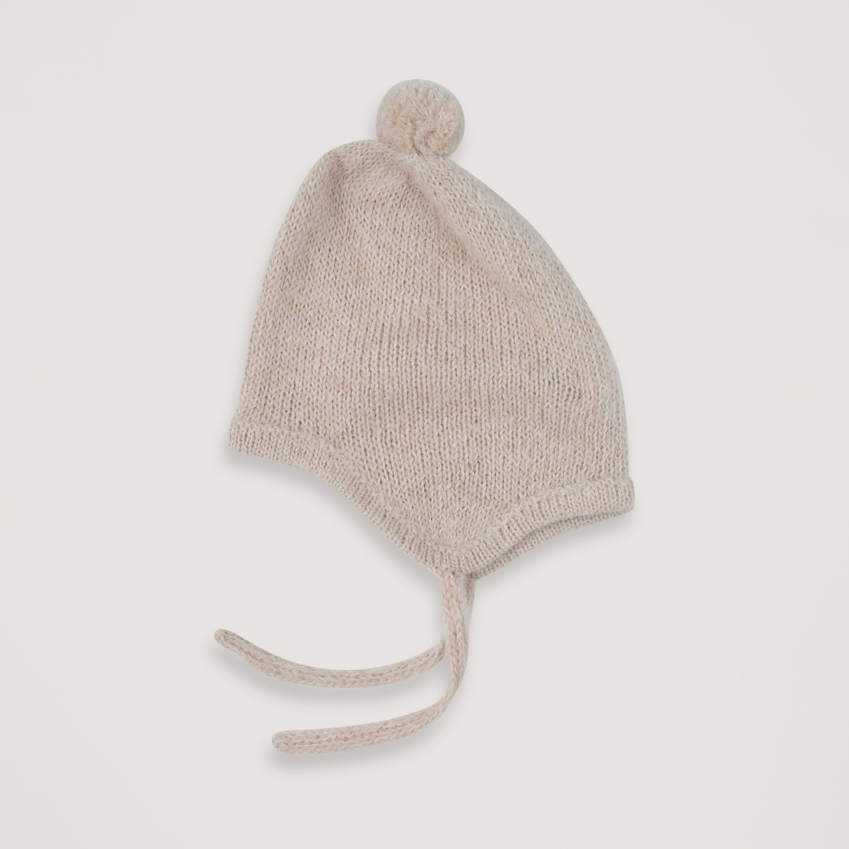 Beige knit baby bonnet with ties and a pompom on a neutral background.