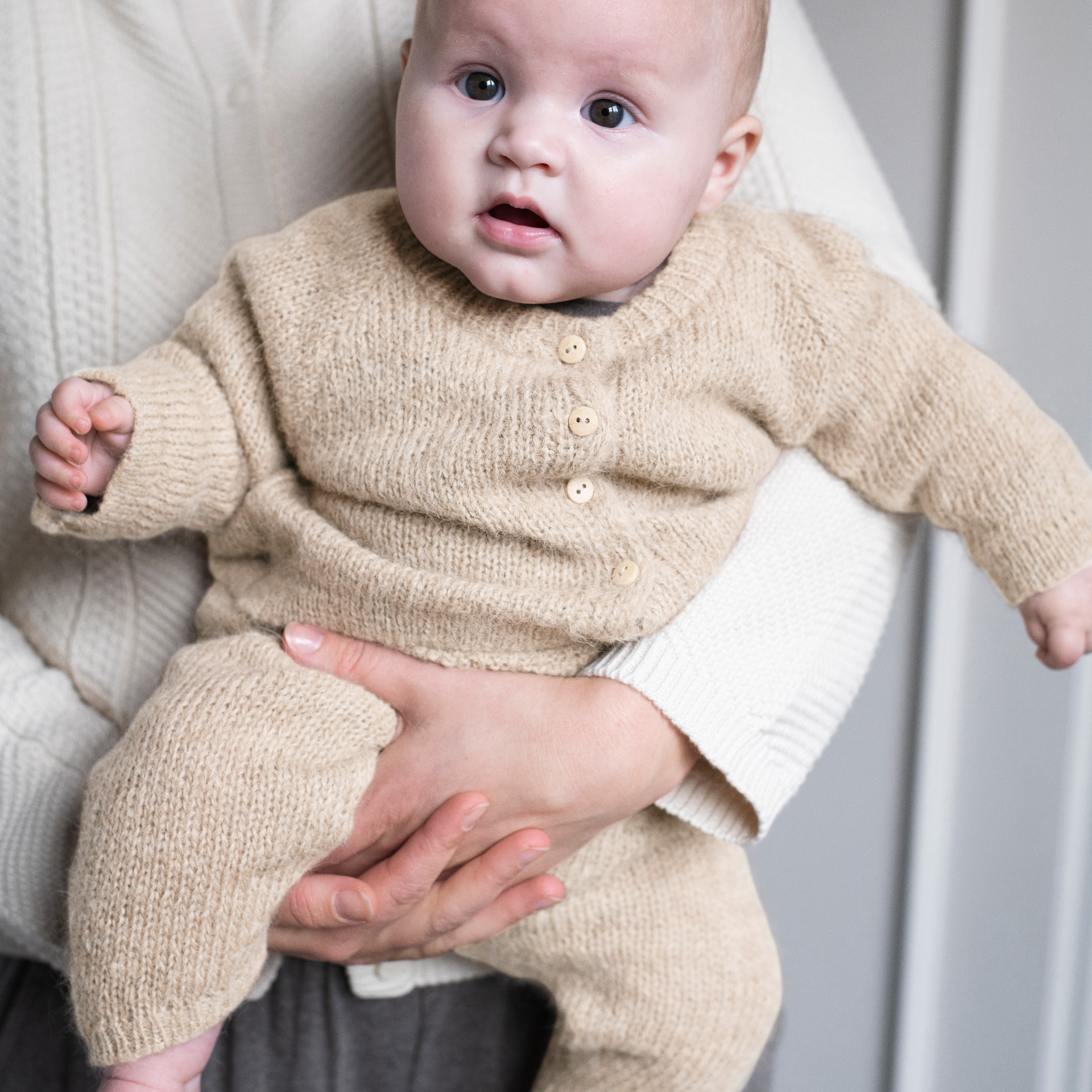 A baby wearing a beige knit cardigan and matching knit pants, held by an adult wearing a cream ribbed sweater. The baby is looking directly at the camera with a neutral gray background.