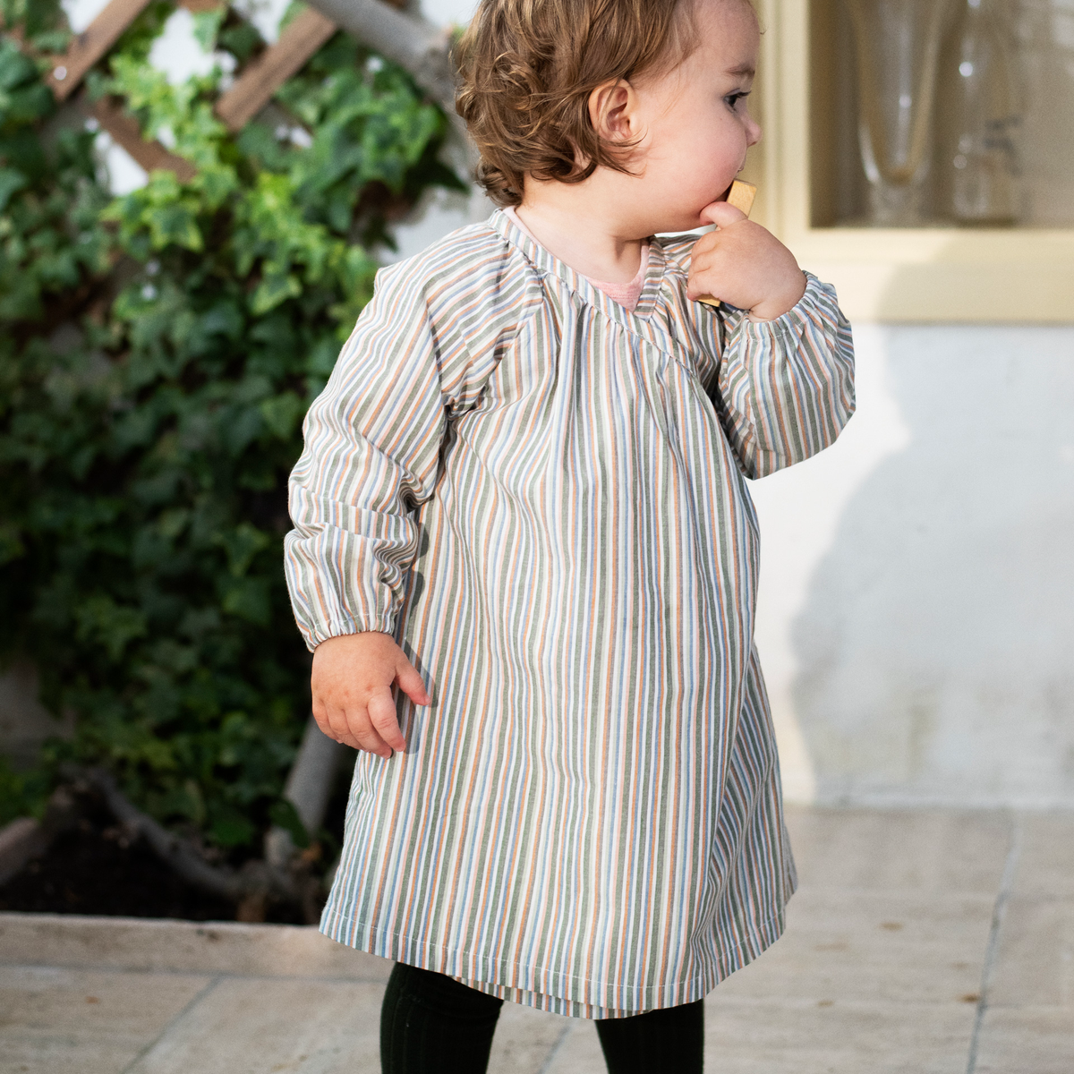 A toddler wearing a long-sleeve baby tunic dress with vertical stripes in beige, blue, and orange, standing outdoors on a patio.