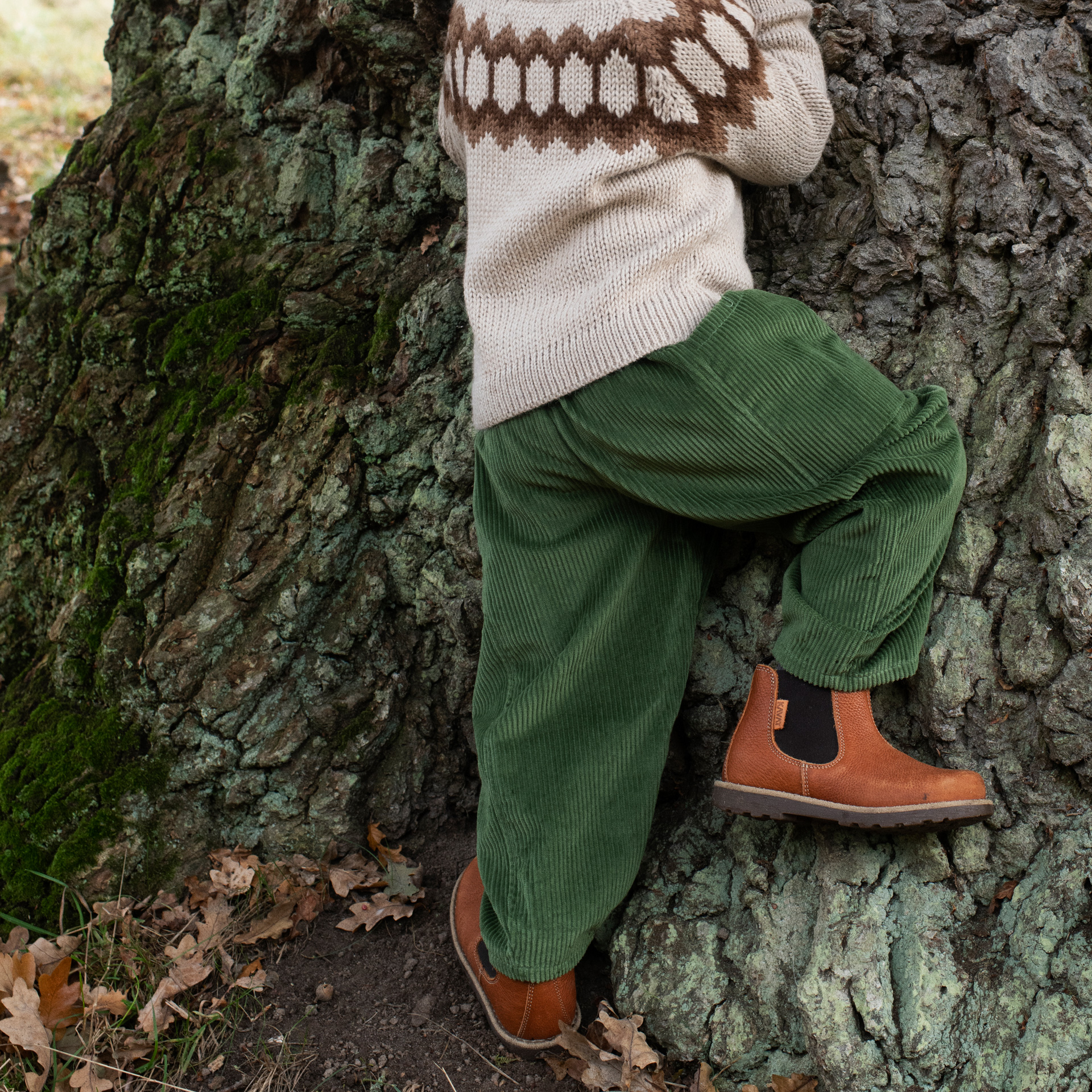 A toddler stands against a large tree trunk outdoors, wearing a cream knit sweater with a brown geometric pattern on the shoulders and sleeves, green corduroy pants, and tan Chelsea-style boots.